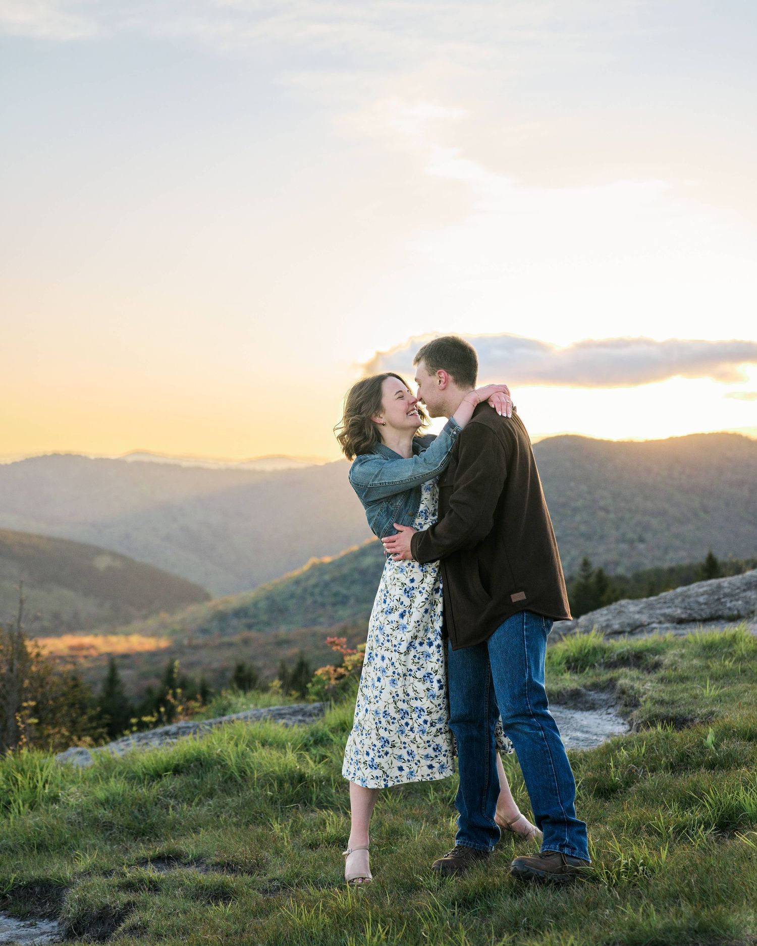 Couple embracing at golden hour sunset with scenic mountain range backdrop on rocky overlook.