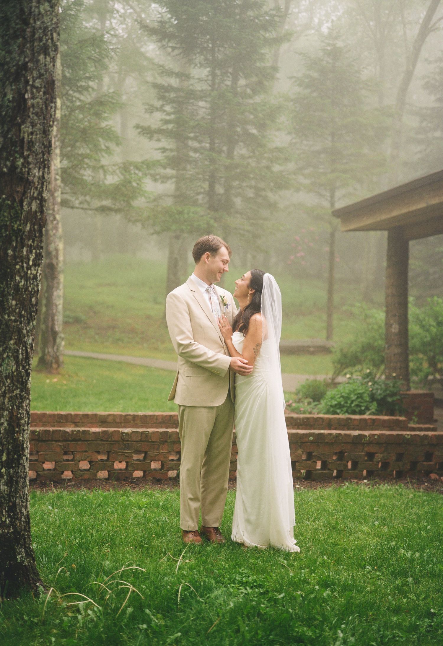 Bride and groom embrace in a misty forest garden setting, she in white gown with veil, he in beige suit.