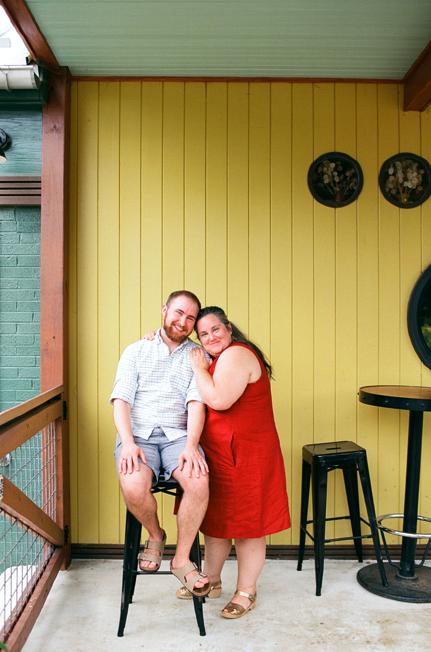A smiling couple poses together against a yellow wall, woman in red dress leaning on seated man in shorts.