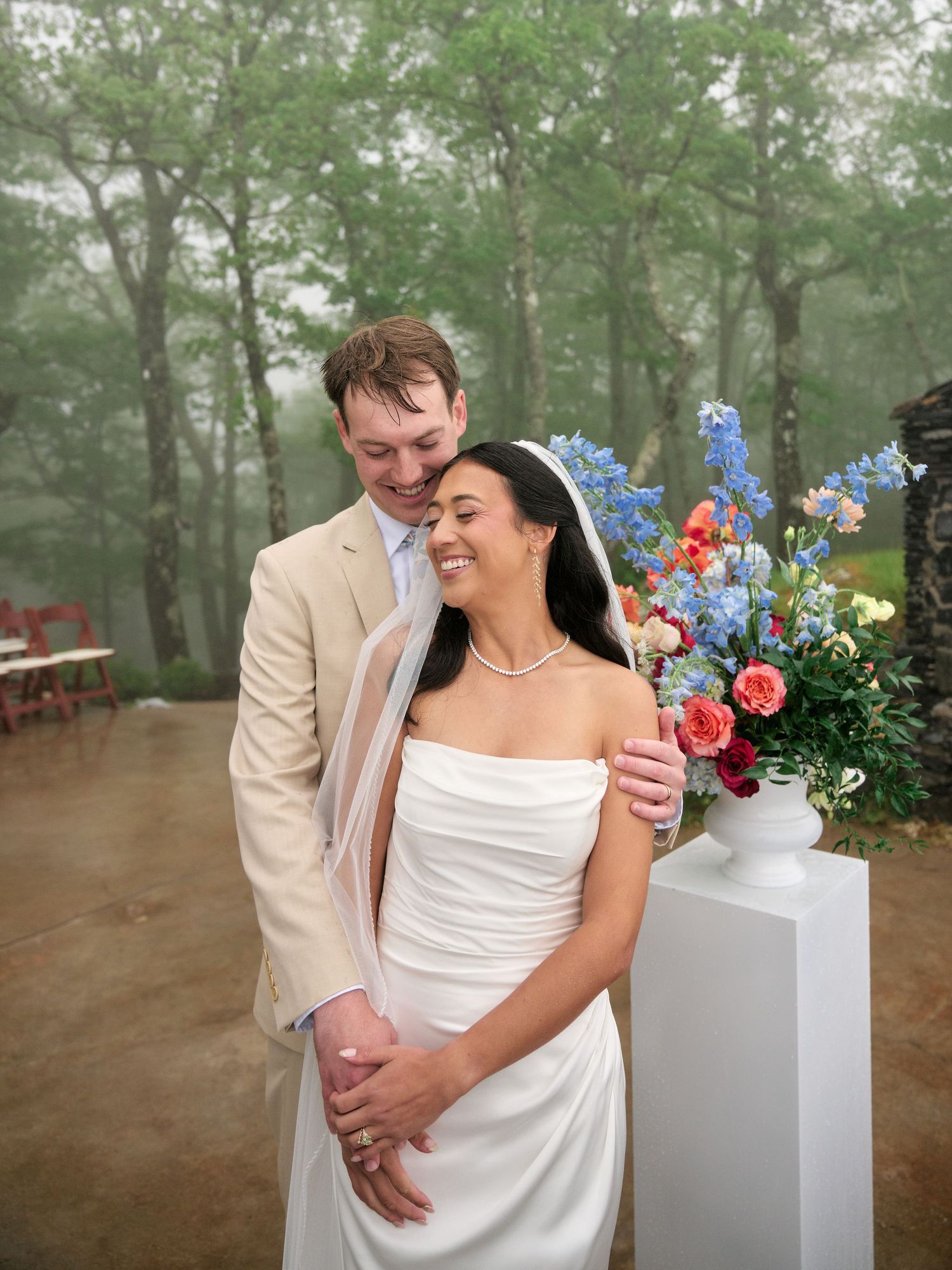 A couple shares a joyful moment at their misty outdoor wedding ceremony, bride in white gown, groom in tan suit.