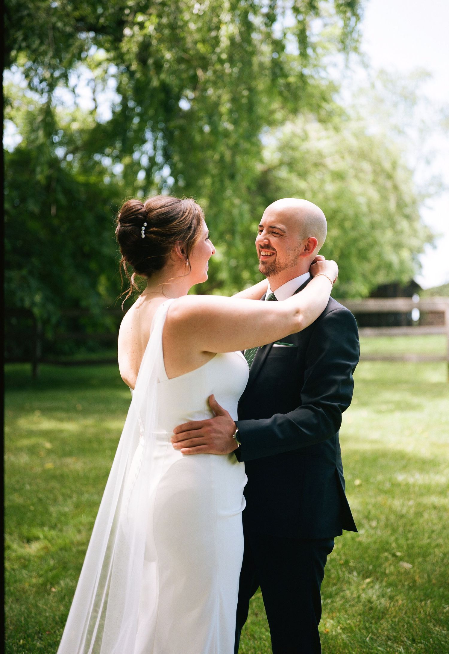 A bride in a white gown and groom in a black suit share a tender dance outdoors on a sunny day.