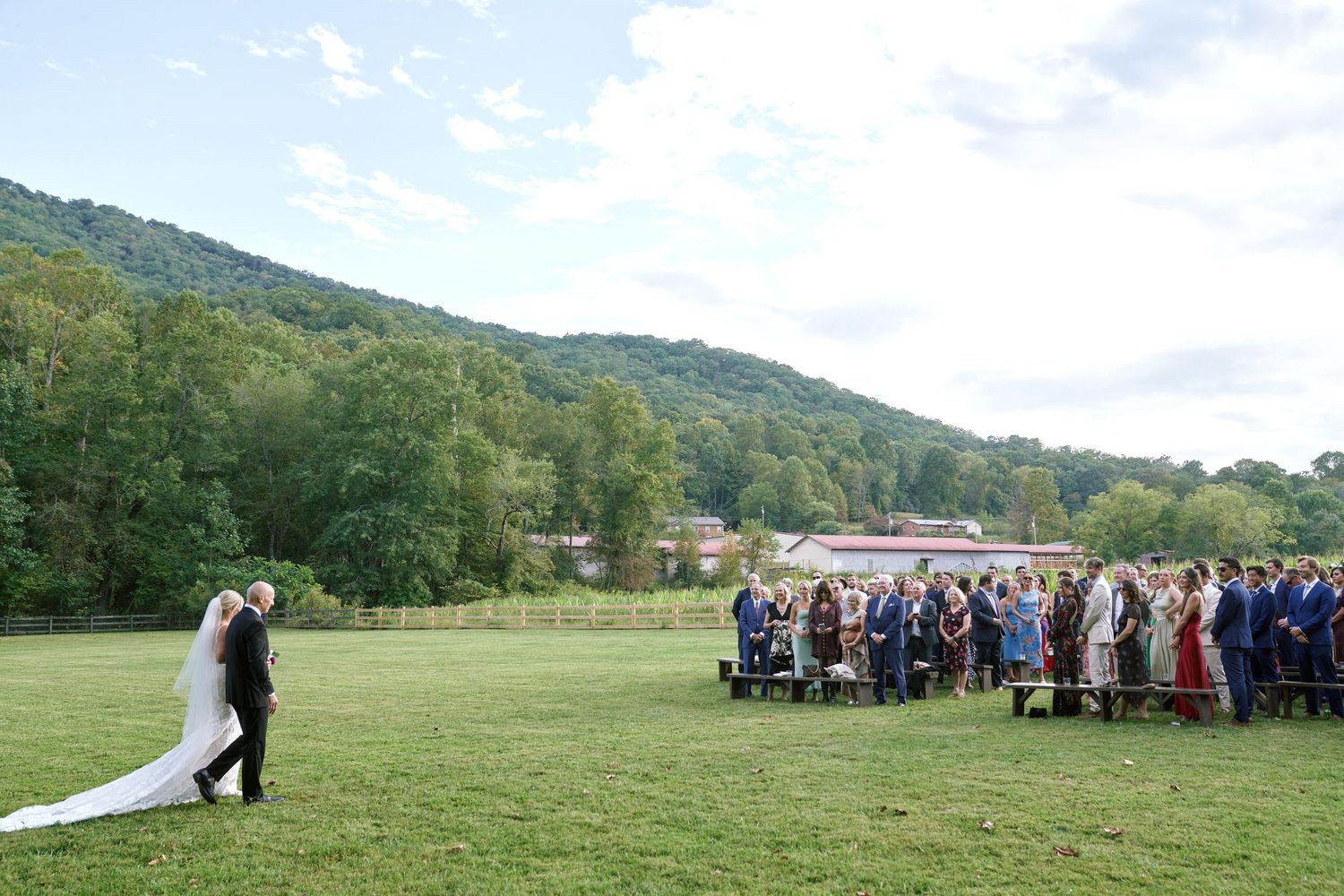 A panoramic view of a green field wedding ceremony setup with chairs and guests gathered under blue skies.