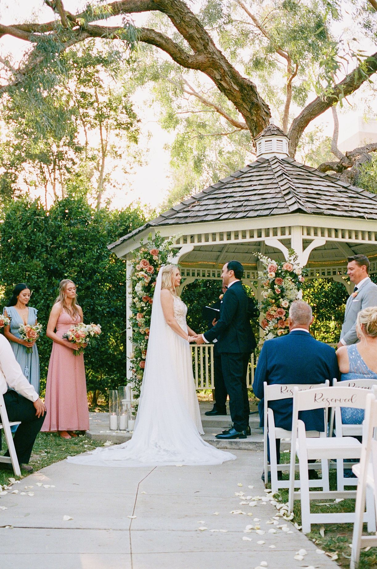 A wedding ceremony takes place under a white gazebo surrounded by trees during a beautiful outdoor celebration.