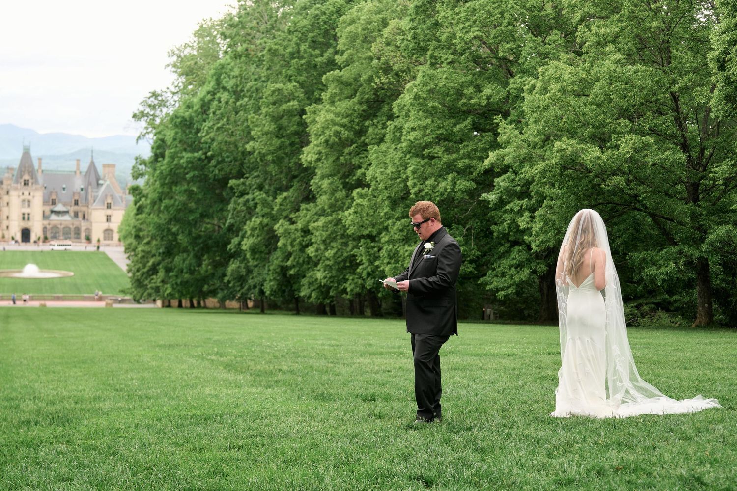 A wedding couple stands on a lush green lawn with a grand castle and trees in the background during their first look moment.