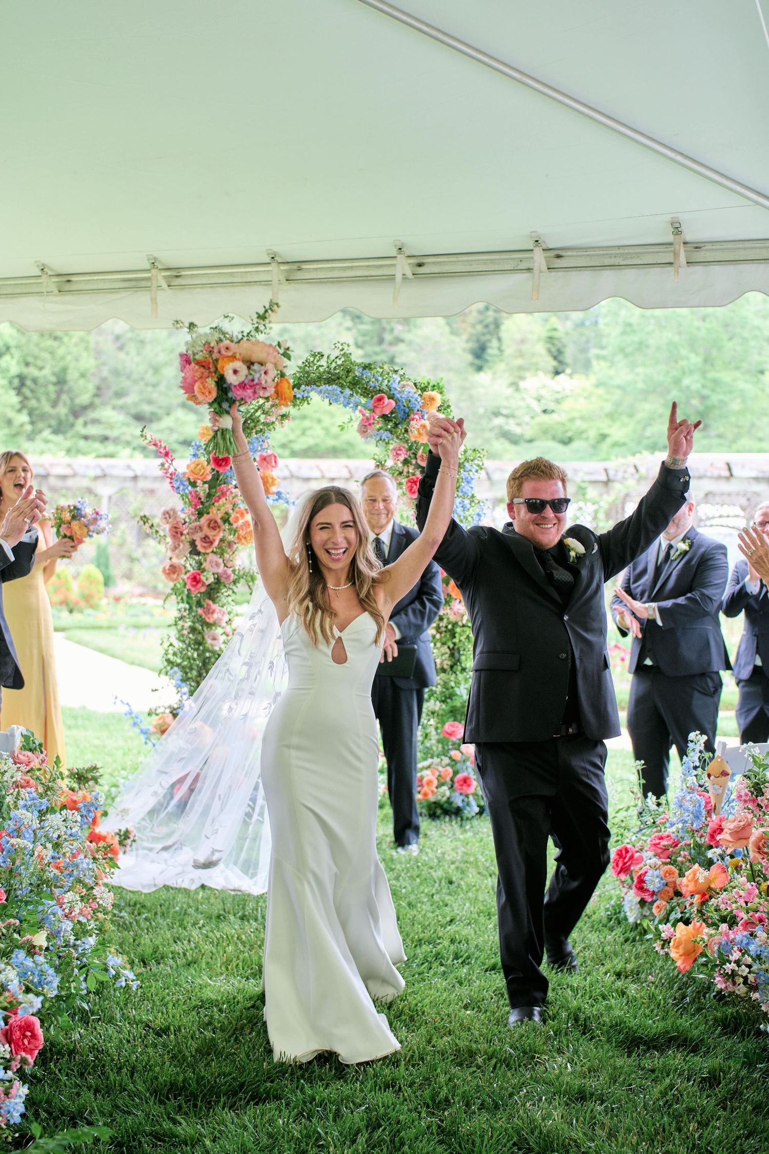 Wedding party celebrates with joyful raised arms under white tent on grassy lawn with vibrant floral arrangements.