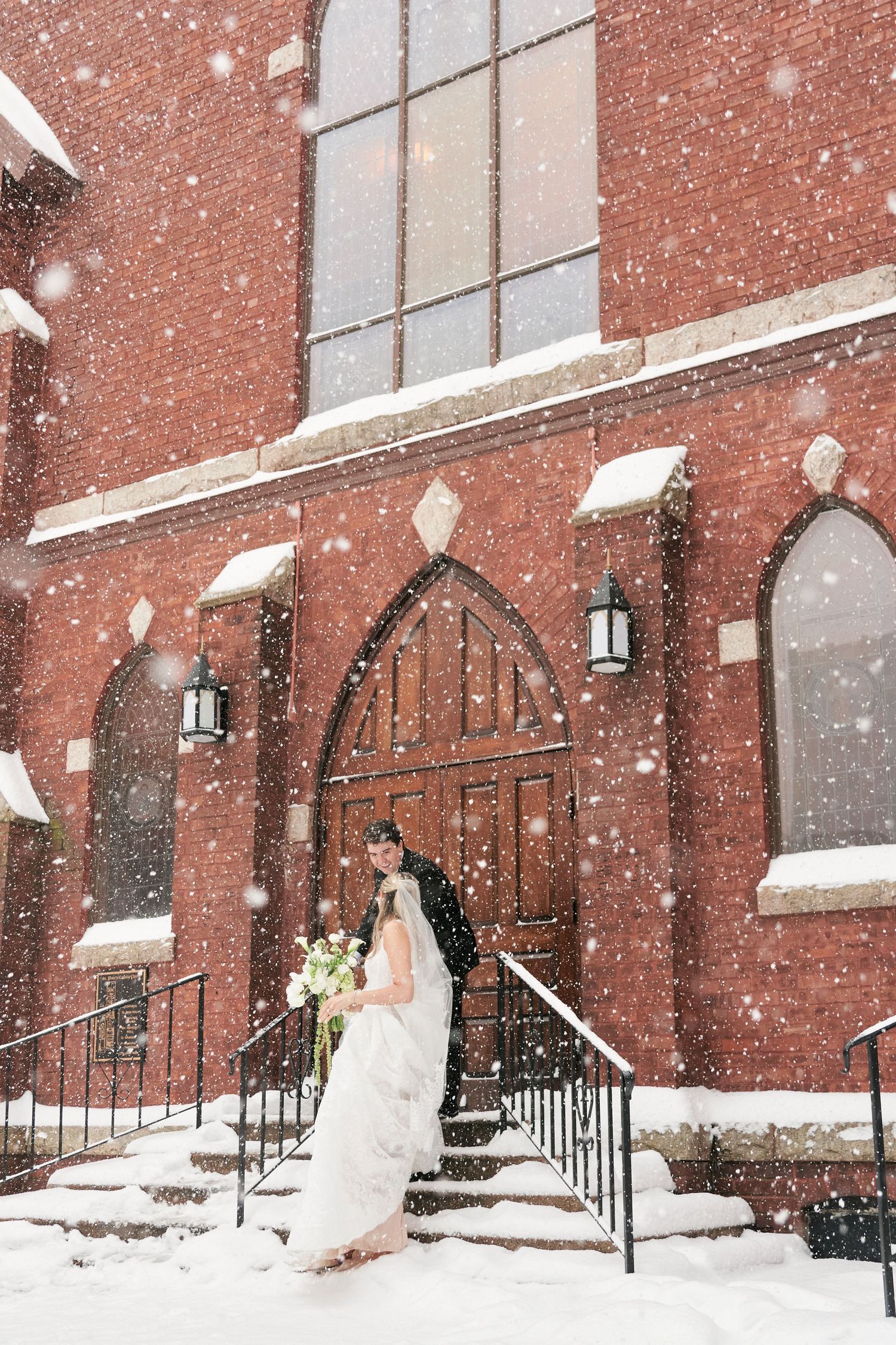 Bride and groom stand on snowy church steps during a winter wedding, with red brick Gothic architecture and falling snow.