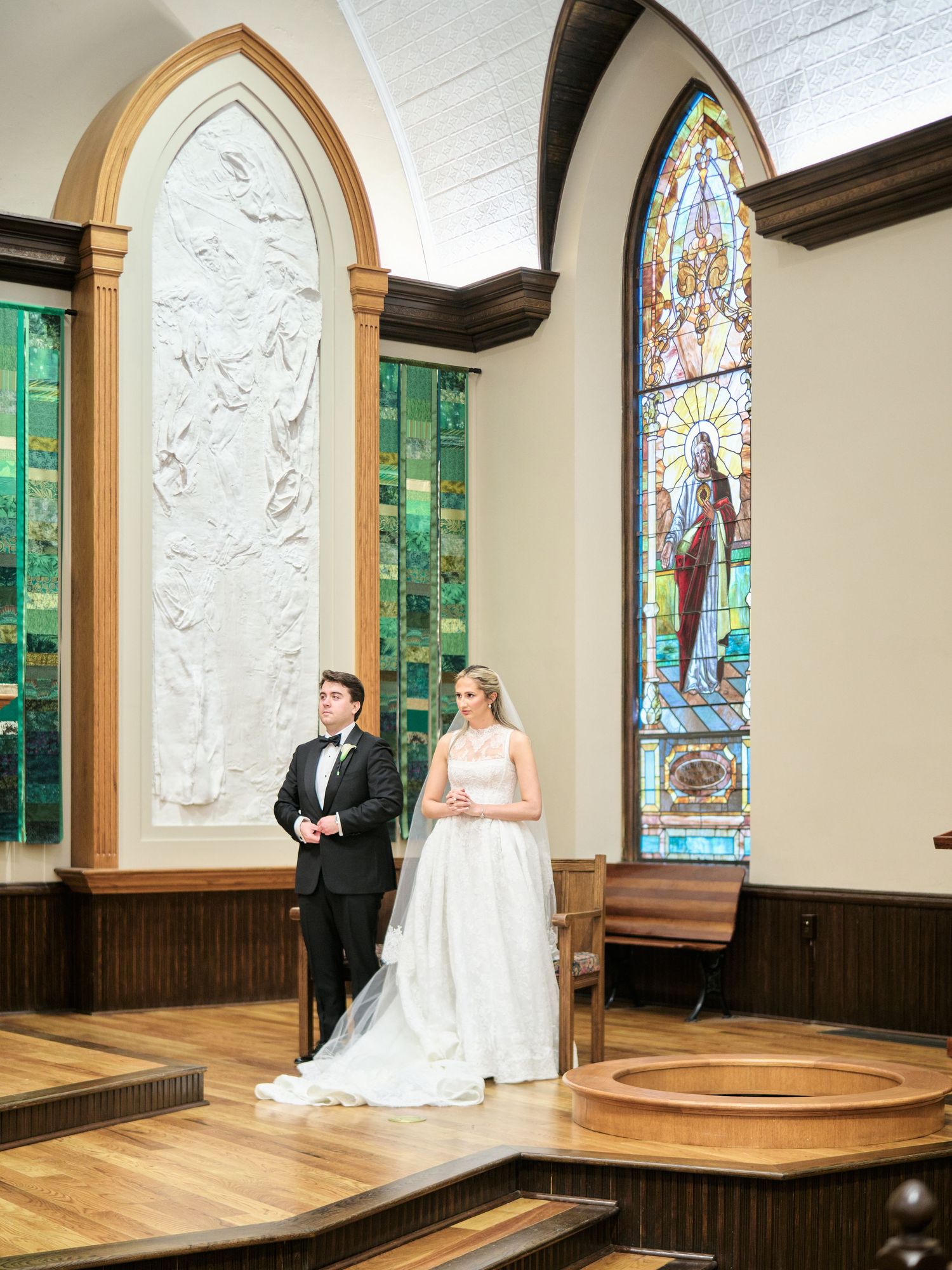 Bride and groom stand at church altar with stained glass windows and white relief sculpture during wedding ceremony.