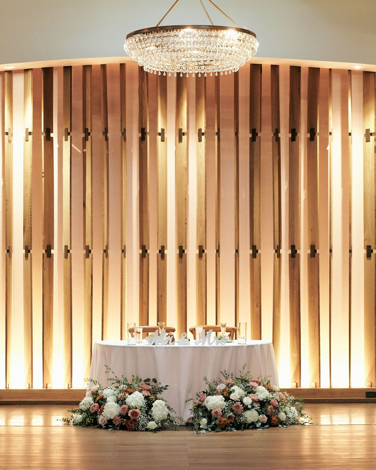 Elegant wedding sweetheart table with white floral arrangements, wood slat backdrop, and crystal chandelier.