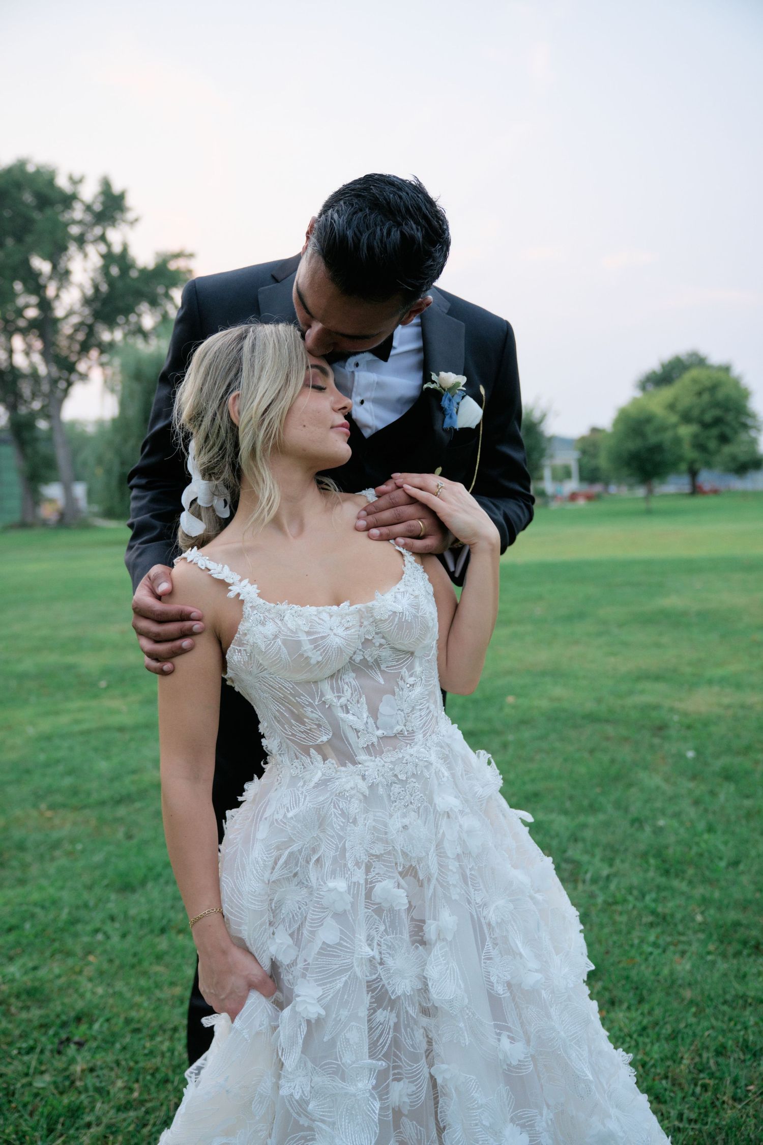 A romantic wedding photograph taken outdoors on a grassy lawn showing an embracing couple in formal wedding attire.