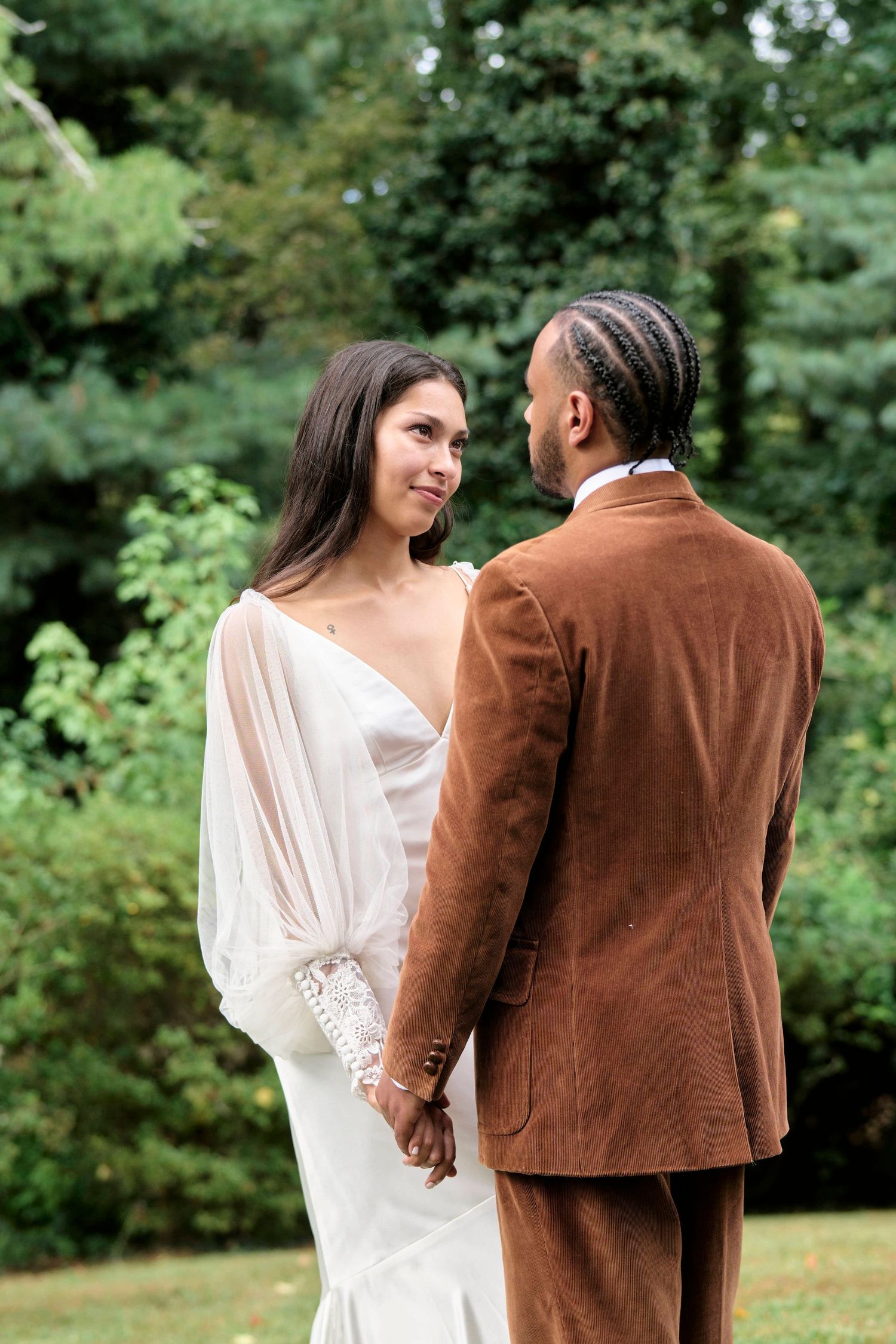 A couple in wedding attire holds hands while looking at each other in a garden.