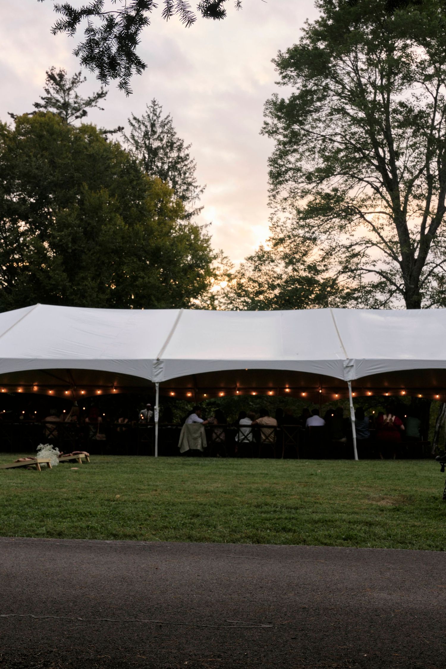A large white reception tent with string lights glows at sunset on a grassy lawn.