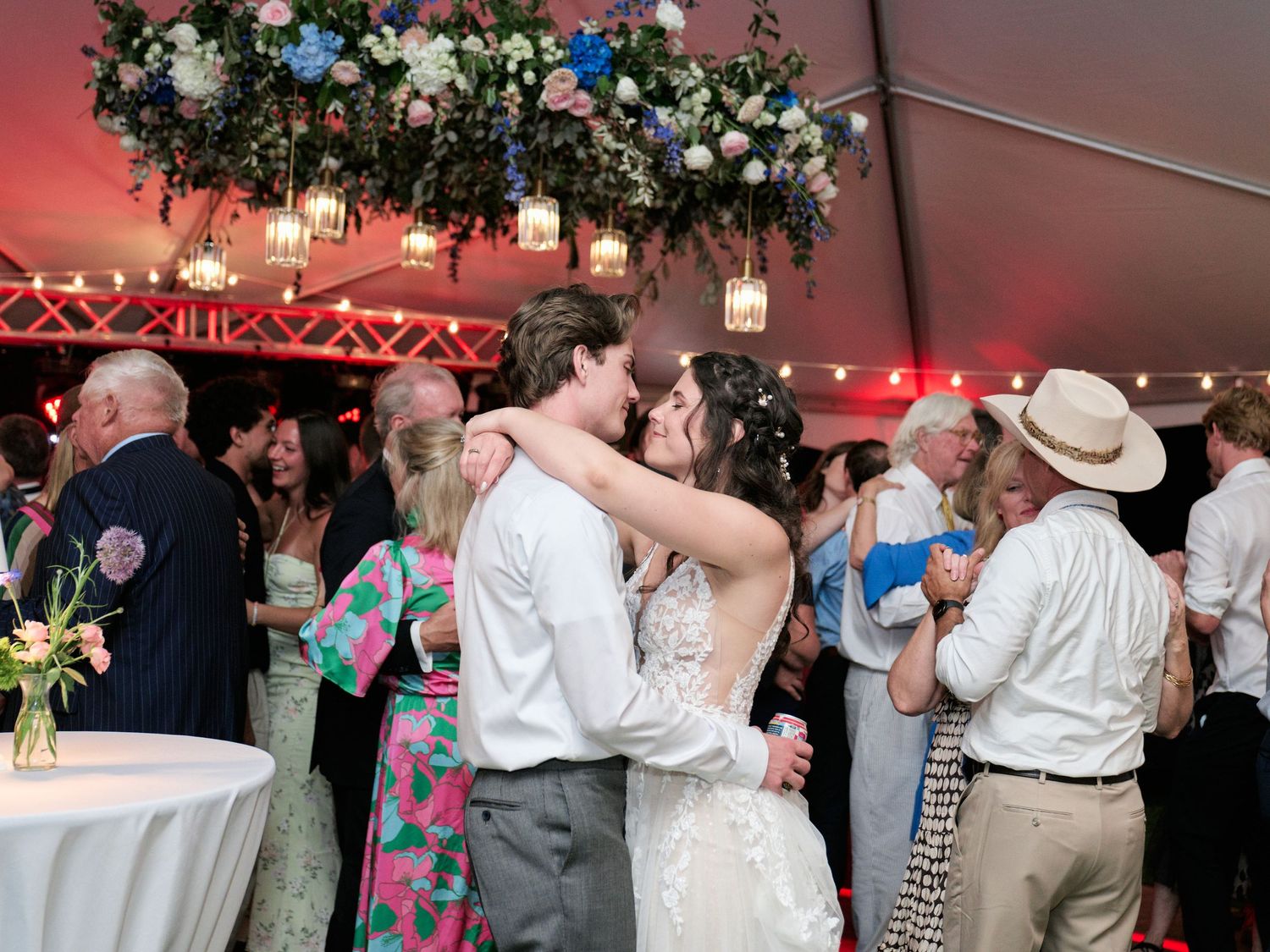 Wedding reception with couples dancing under floral decor and lanterns in a tent with red lighting.