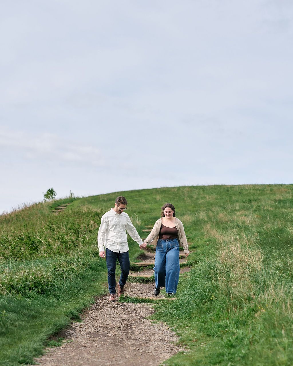 A couple walks hand in hand along a grassy hillside path on a cloudy day.