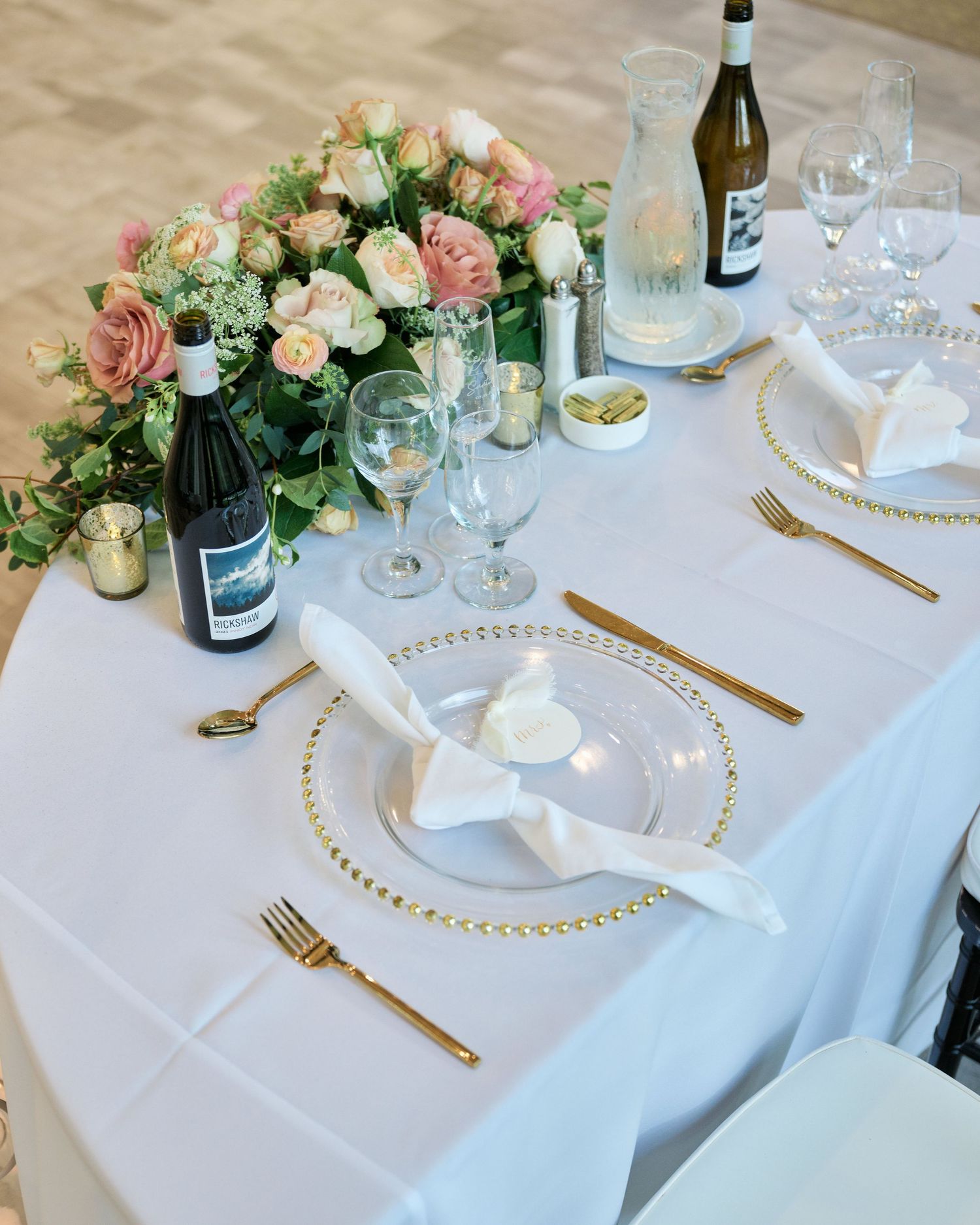 An elegant wedding table setting with pink roses, wine glasses, and gold cutlery on white linens.