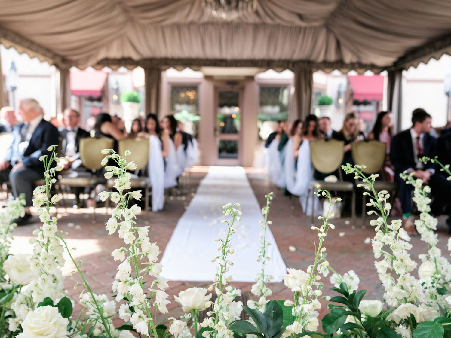 White floral arrangements line a covered walkway with draped fabric ceiling and pink accents at an outdoor event.