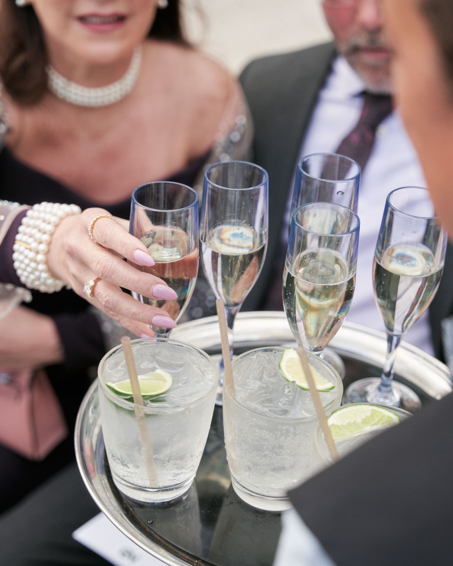 Champagne flutes and cocktail glasses with lime garnishes are arranged on an elegant silver serving tray at a formal event.