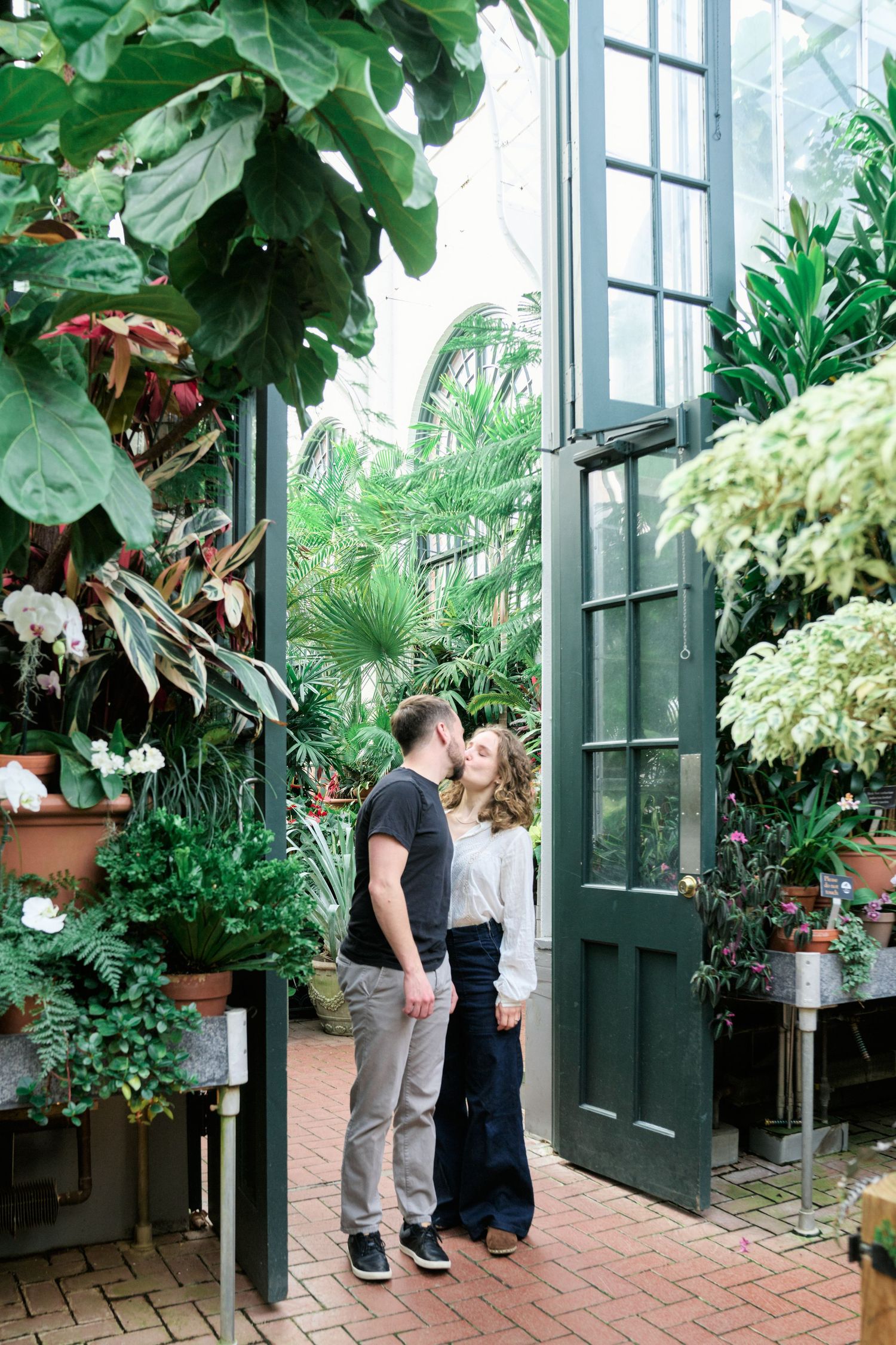 A romantic moment shared in a lush greenhouse filled with tropical plants and brick flooring.