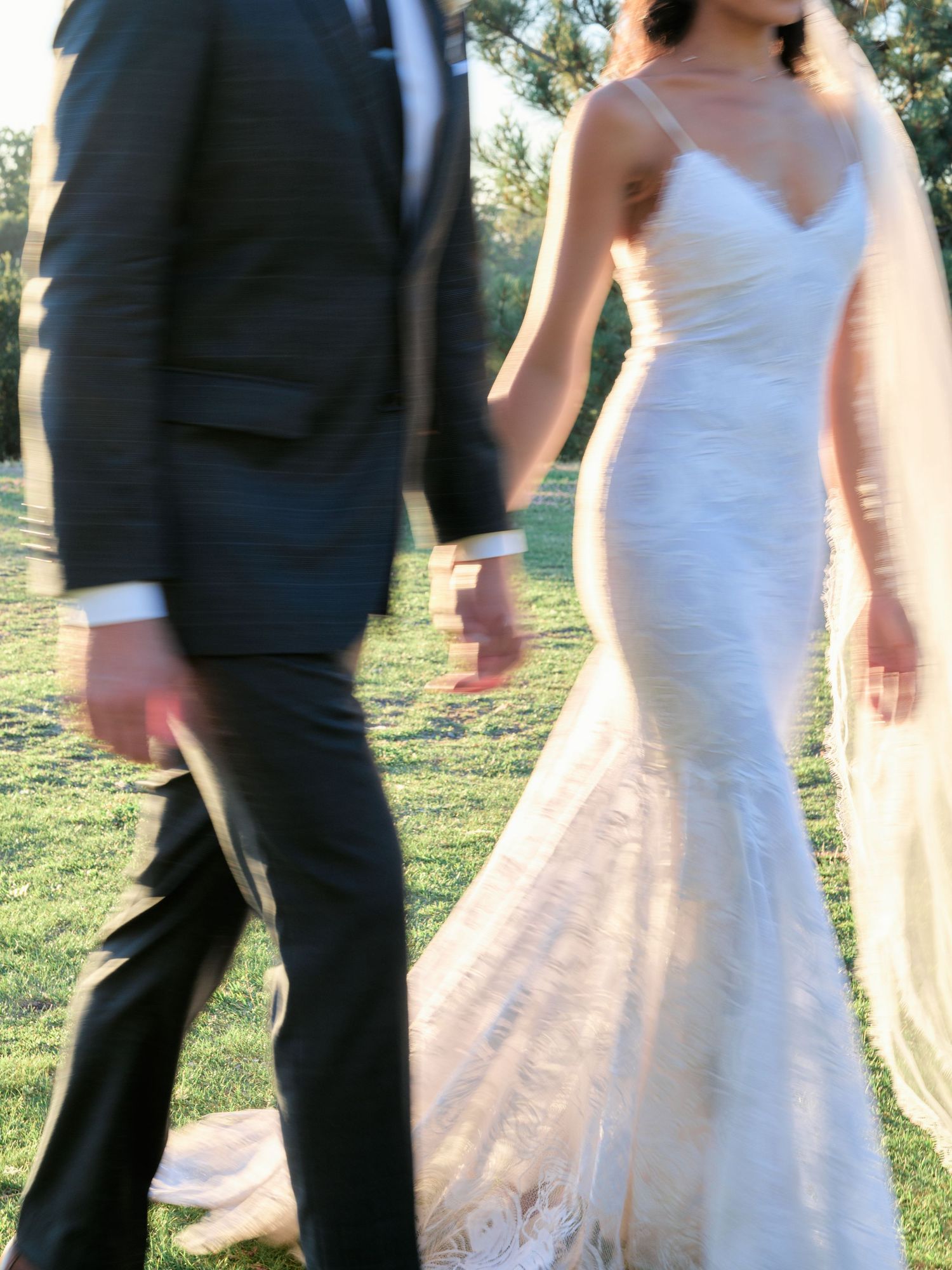 Close-up detail of a fitted wedding dress and black tuxedo as couple walks together.