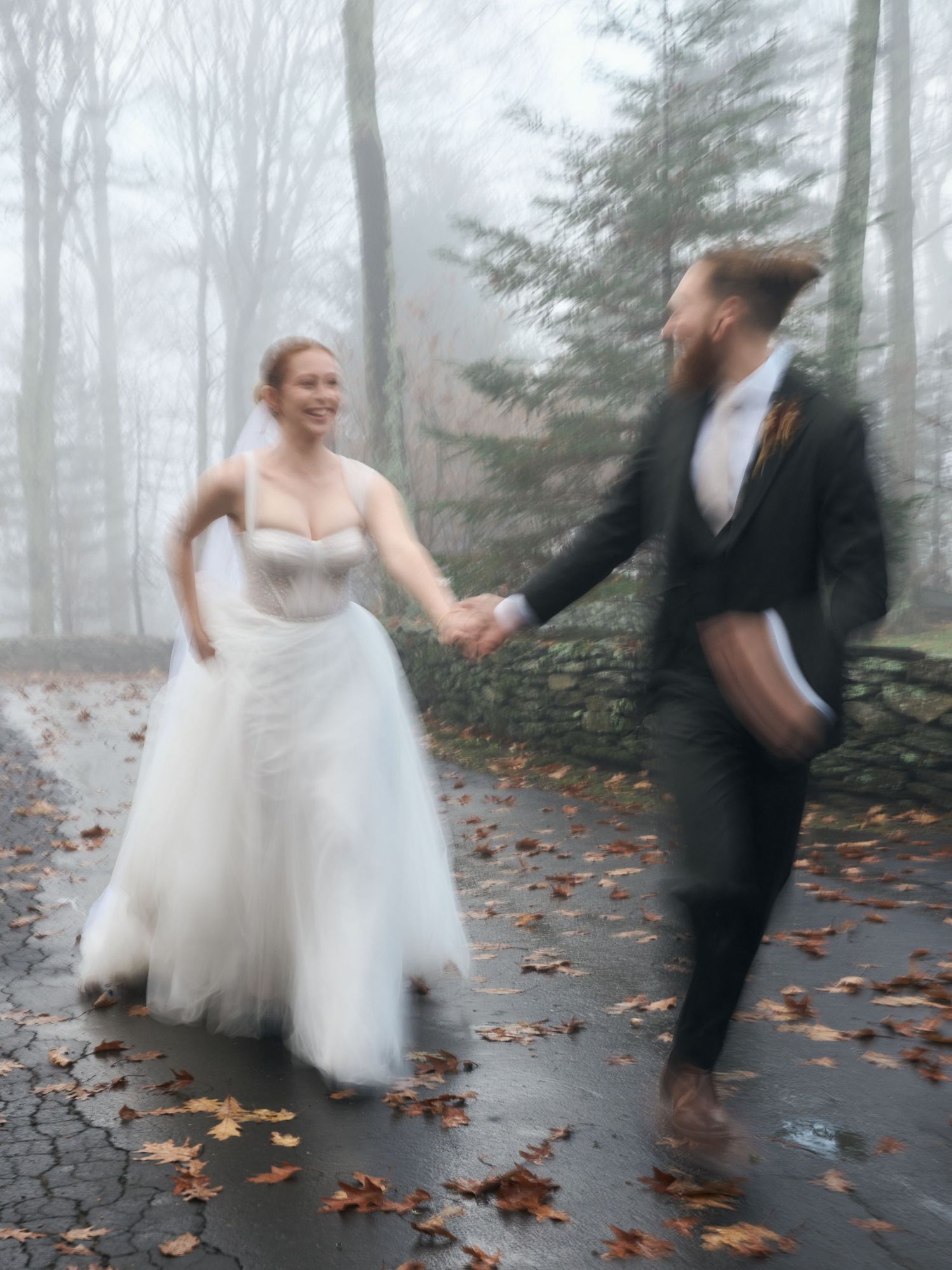 Playful newlyweds dancing together on foggy autumn path covered with fallen leaves.