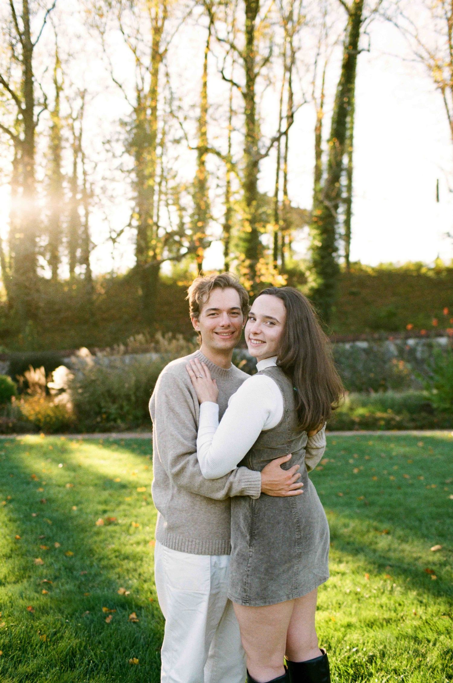 A couple embraces warmly in a sunlit garden with tall trees and green grass in the background.