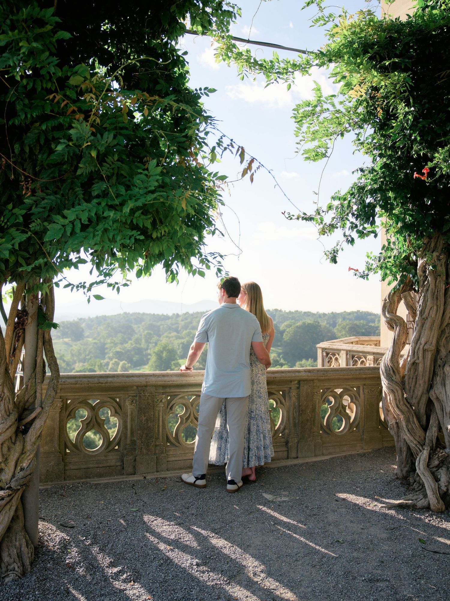People stand at a scenic stone balcony overlooking mountains and trees at a historic estate.