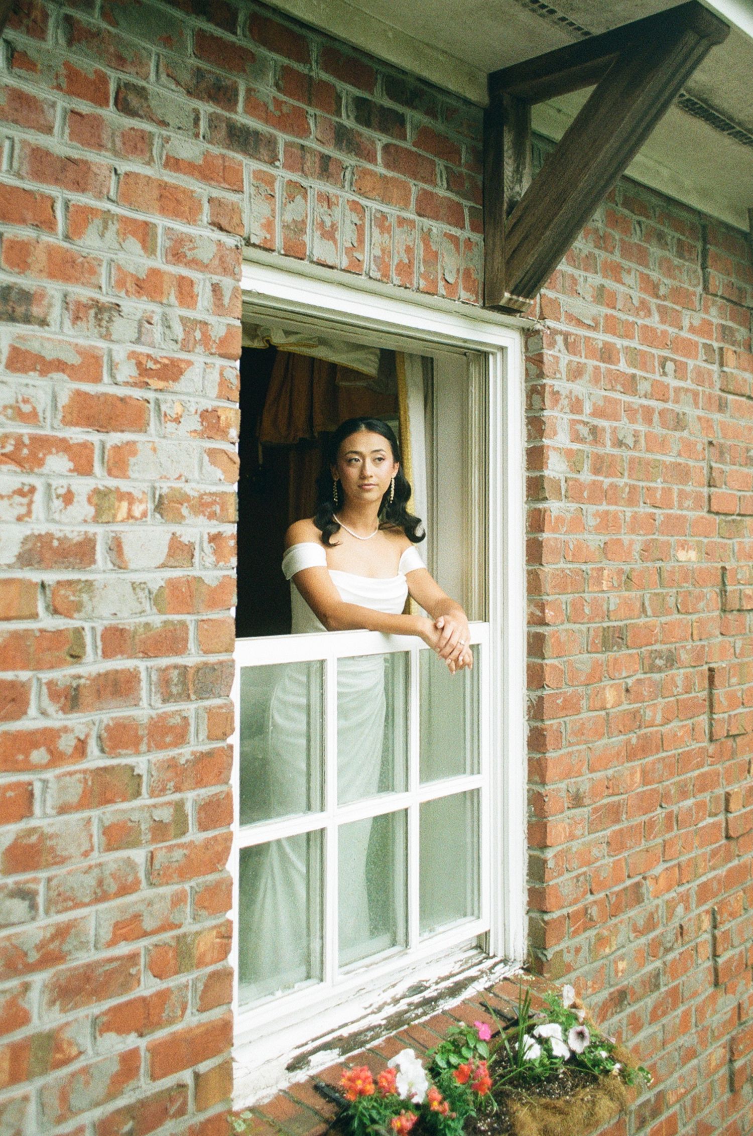 A woman in a white dress leans out of an open window in a red brick building with a flower box below.