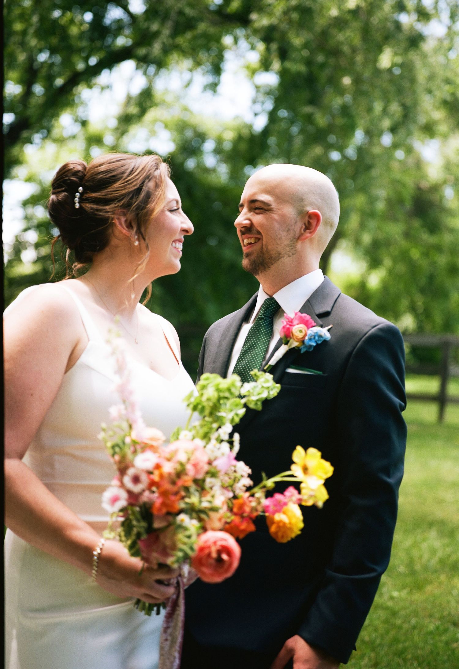 Smiling bride holding colorful bouquet stands with groom in dark suit and floral boutonniere outdoors.