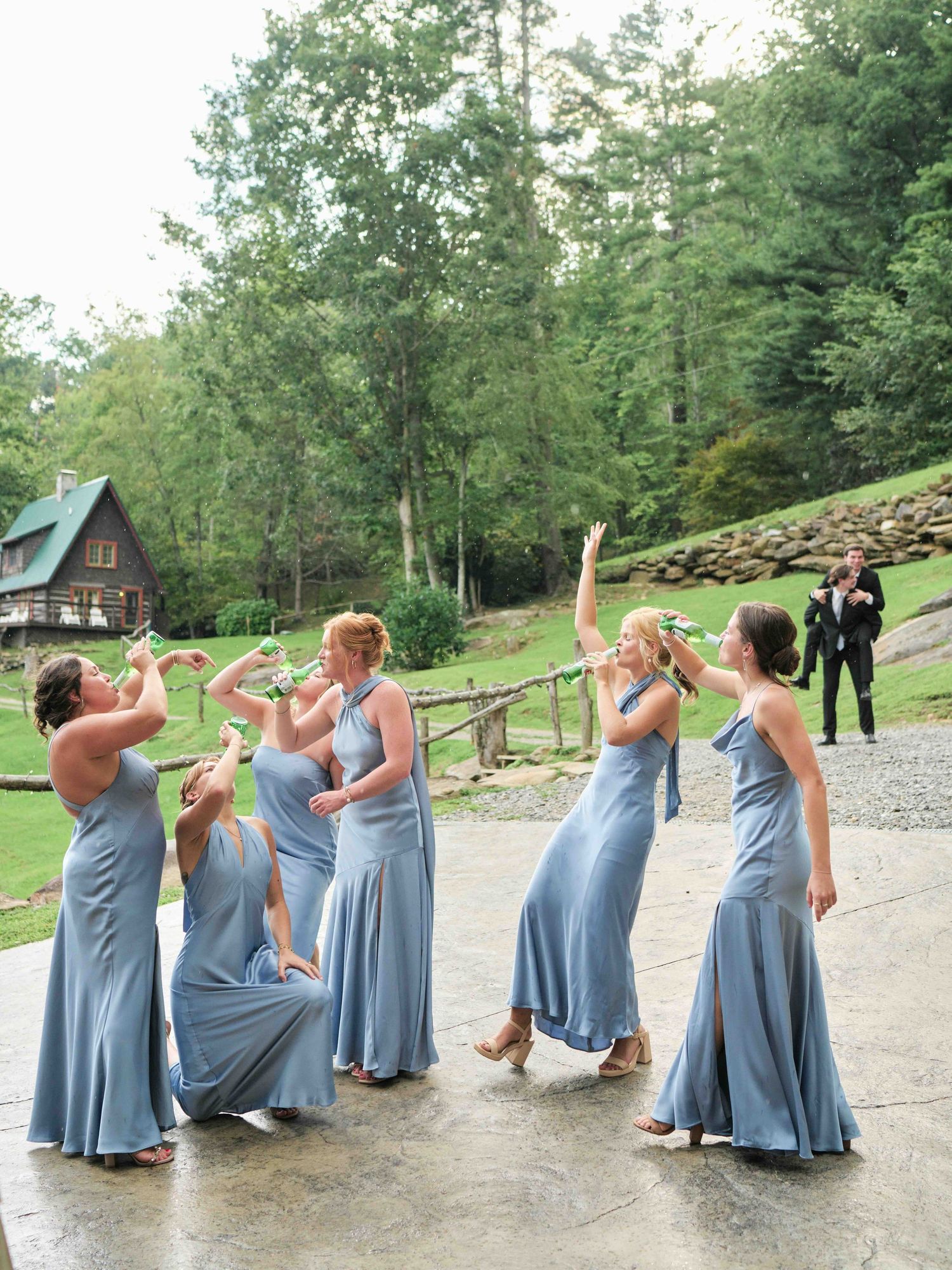 Six bridesmaids in dusty blue gowns celebrate outdoors near a rustic cabin surrounded by lush green trees.