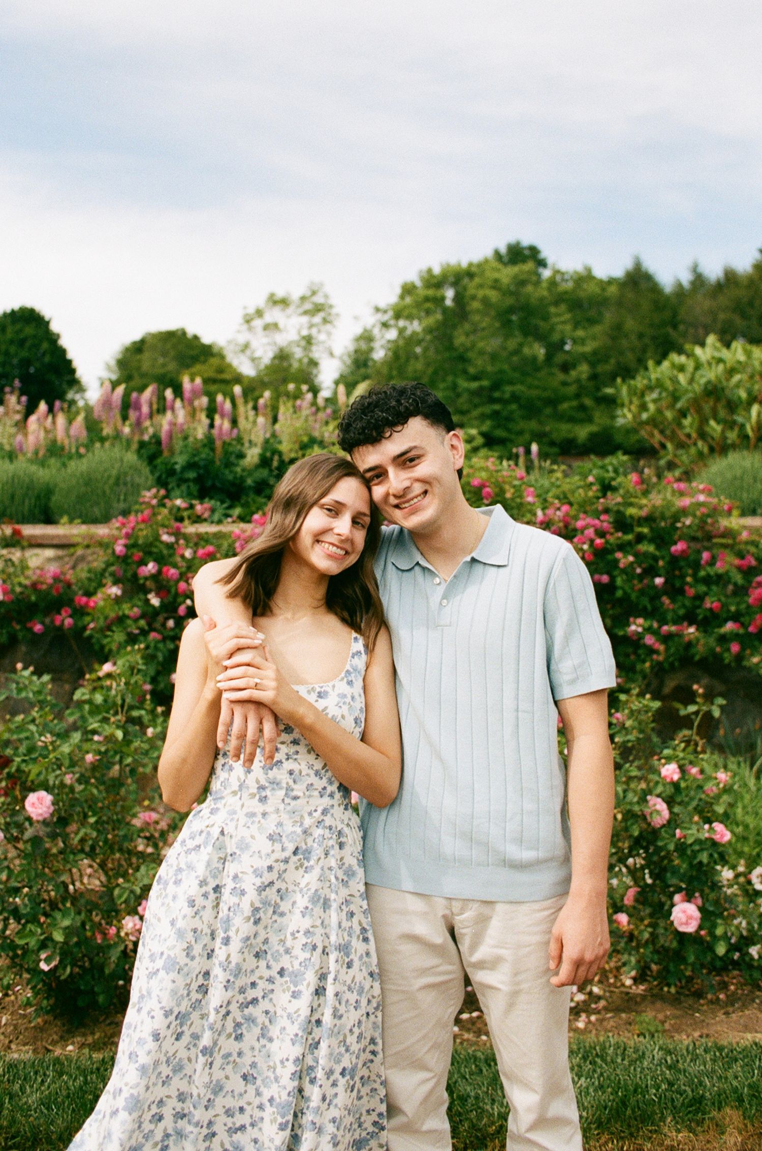 A young couple embraces and smiles in front of blooming rose gardens, woman in floral dress, man in light blue polo shirt.