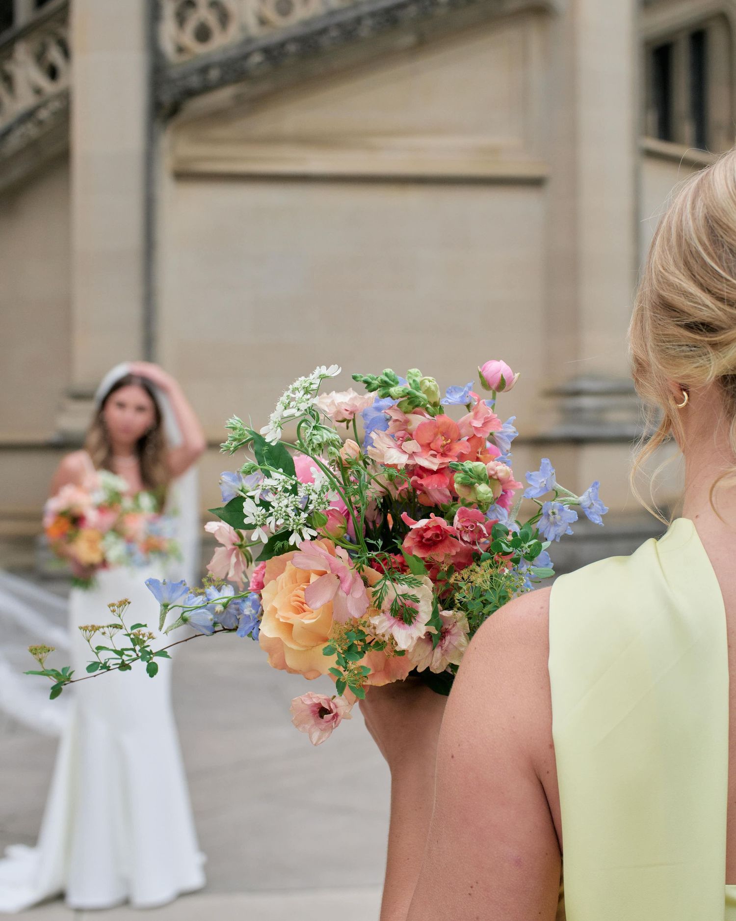 A row of bridesmaids holding colorful floral bouquets with pink, peach and white flowers against a stone building.