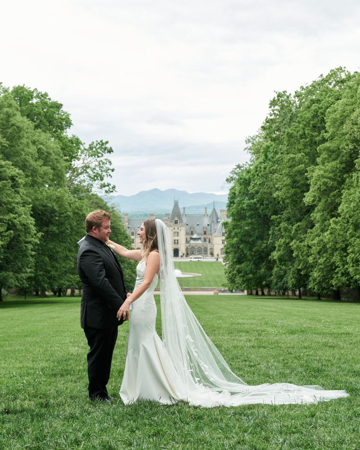 A couple in wedding attire shares an intimate moment on a lush green hilltop with a castle-like building in the distance.