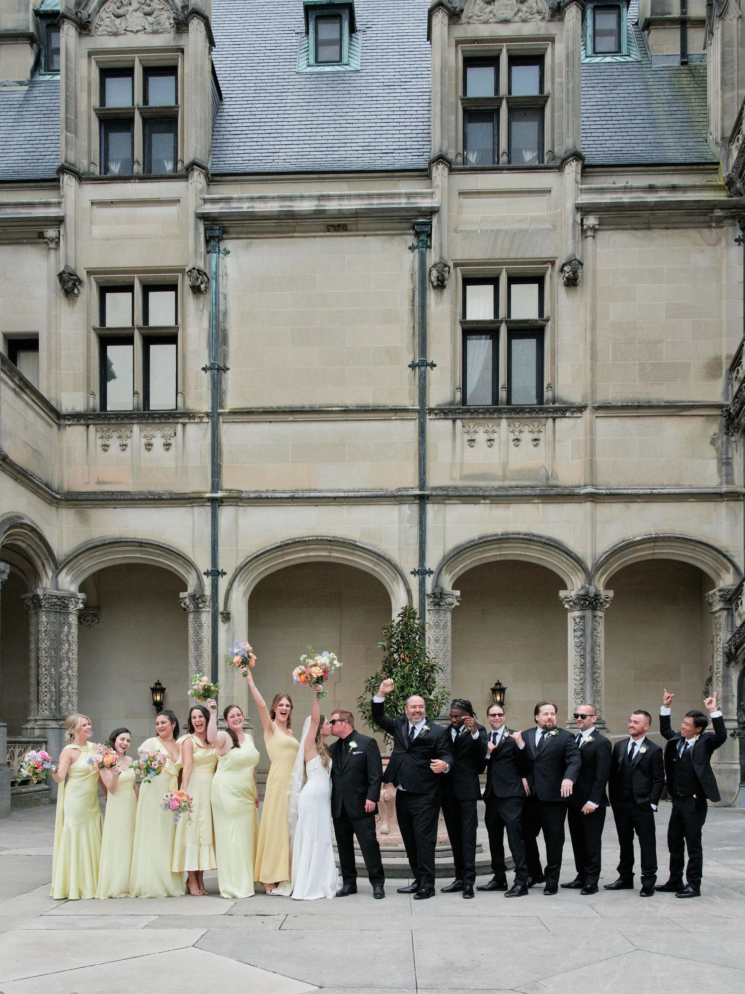 Wedding party members lined up in formal attire along a grand stone building with arched architecture.