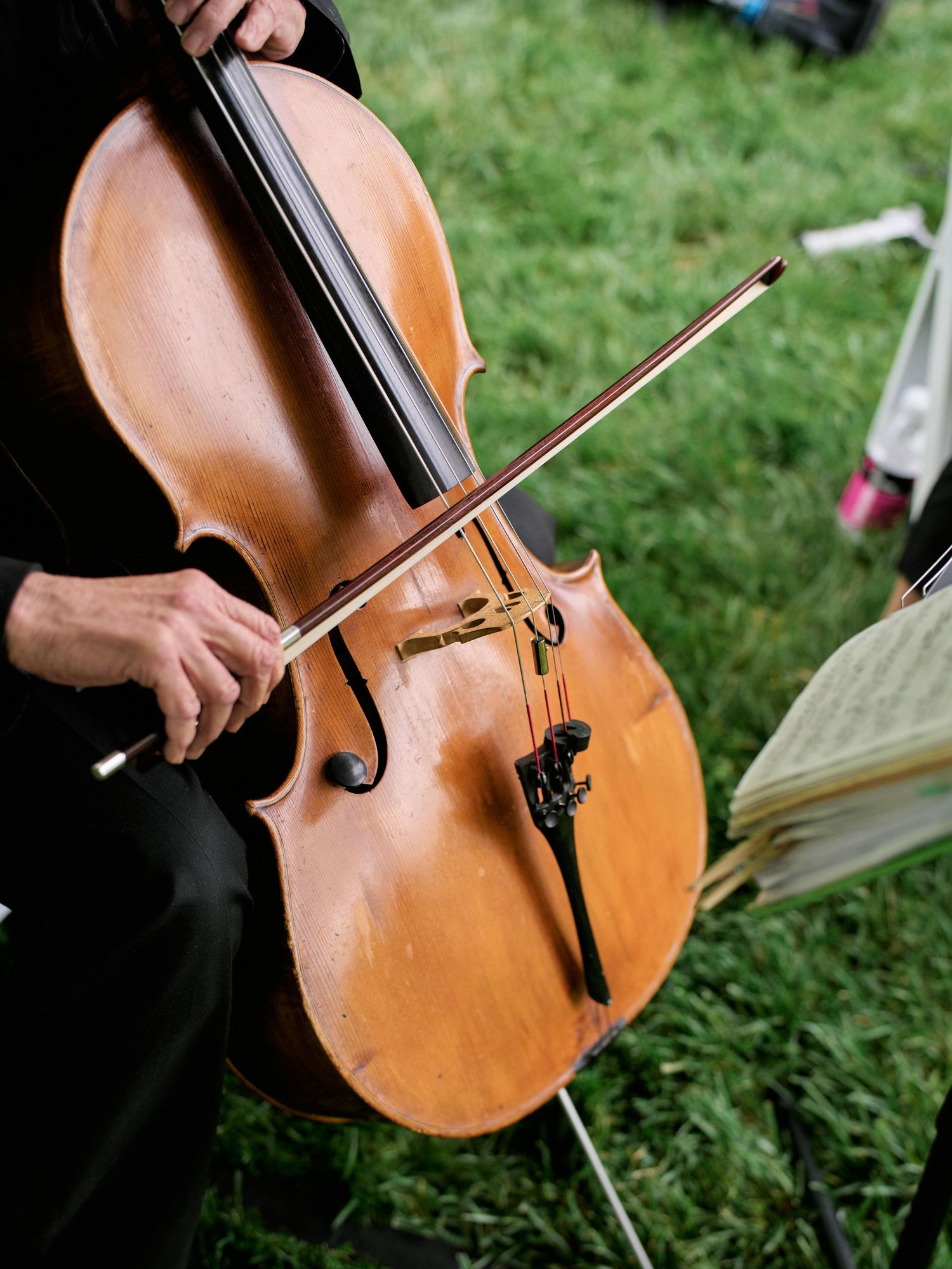 A wooden cello being played outdoors on grass, showing the warm honey-colored wood grain and bow in motion.