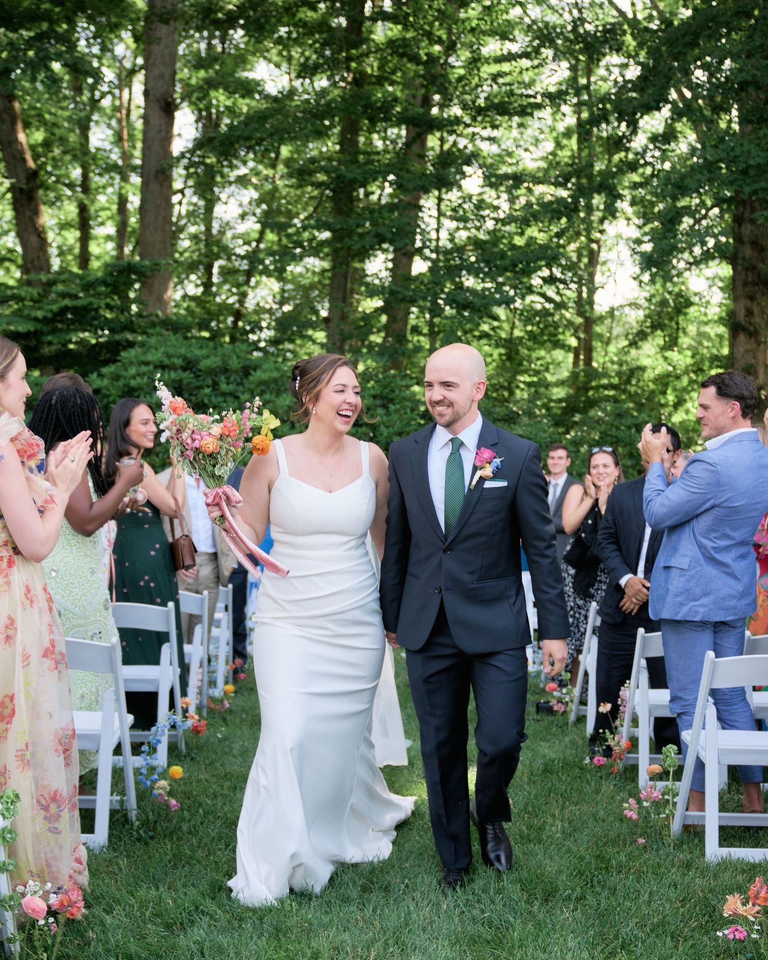 A joyful couple processes down a grassy aisle while wedding guests in colorful attire applaud.