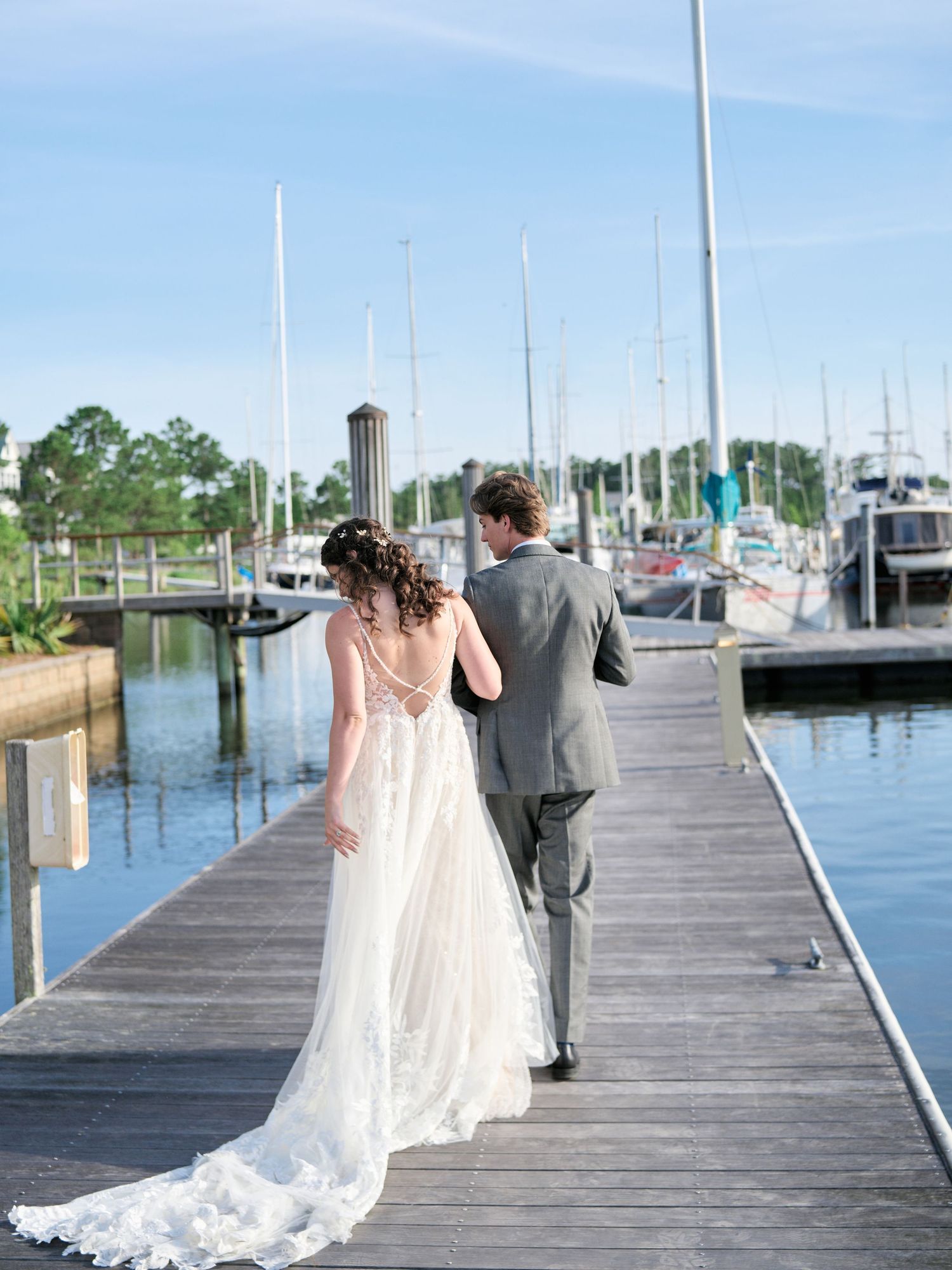 A couple in wedding attire walking together on a marina dock with sailboats and blue sky in the background.