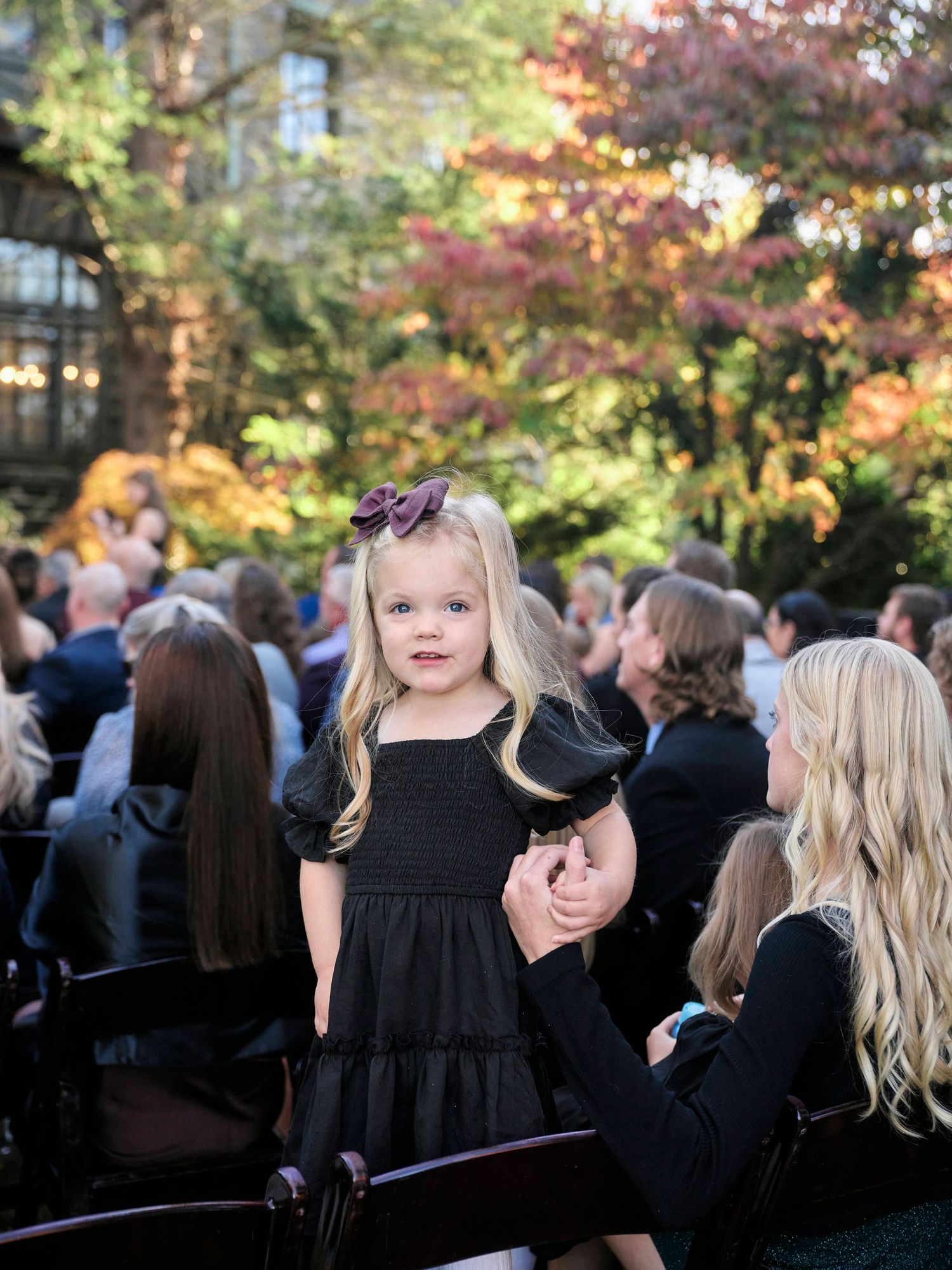 A young attendee in a black dress watches the wedding ceremony among seated guests.
