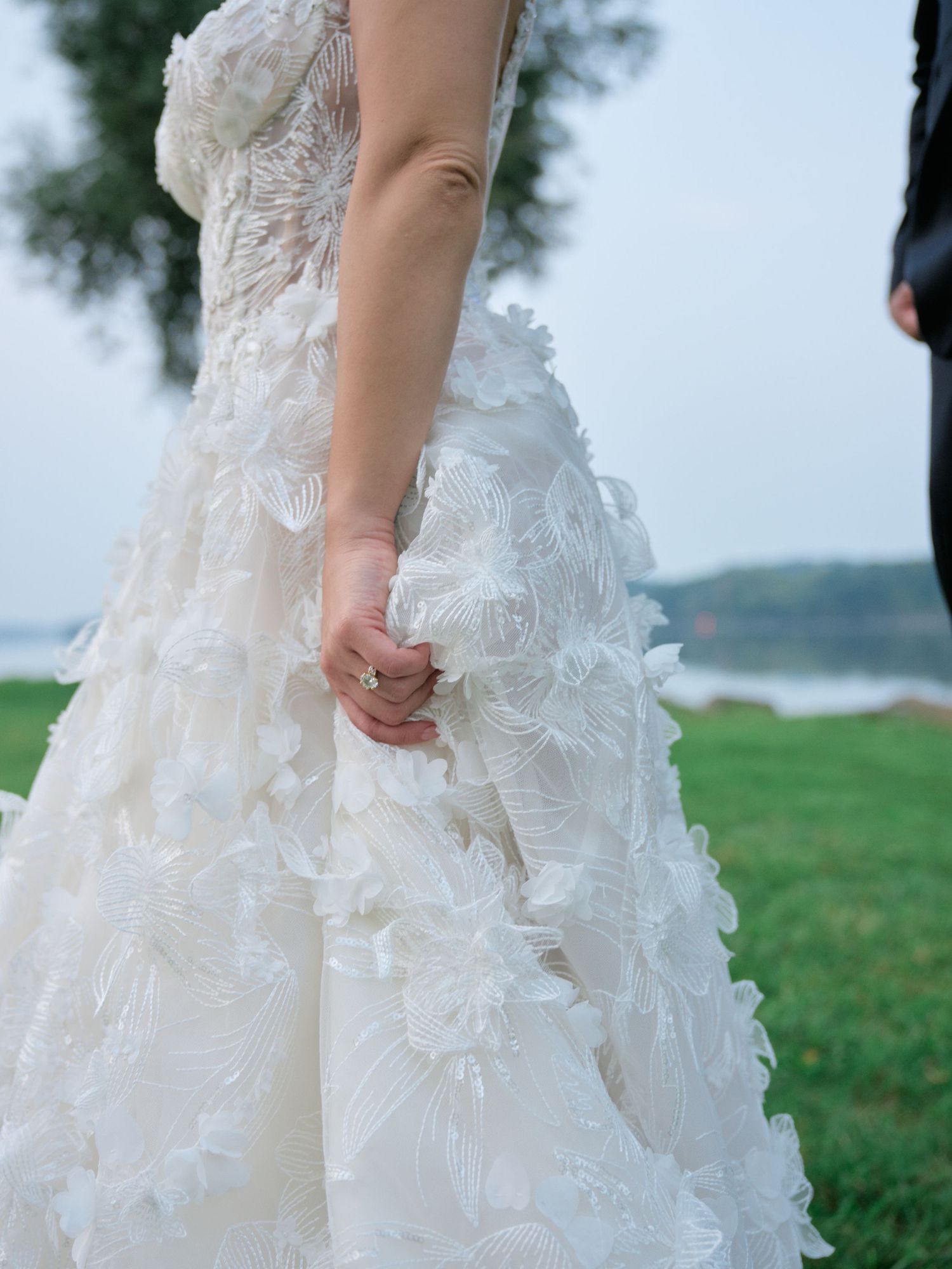 Close-up detail of a white wedding dress with delicate lace and floral appliques against a green outdoor background.