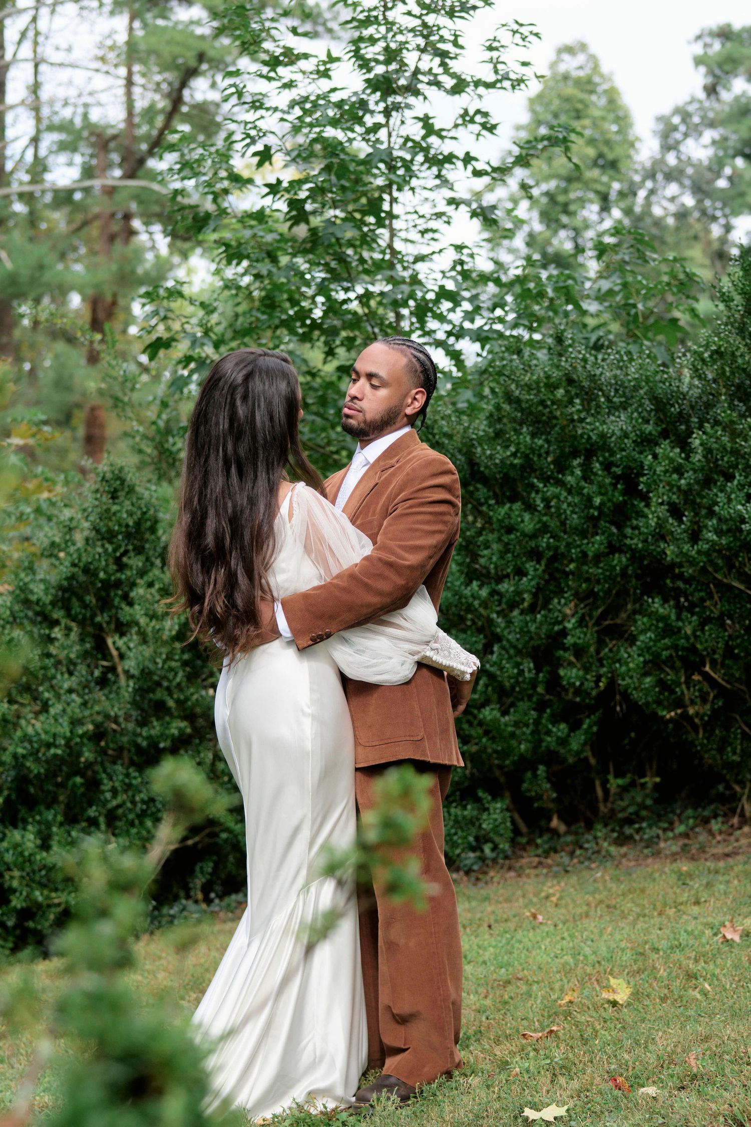 A couple in formal attire embraces in a garden setting with lush greenery.