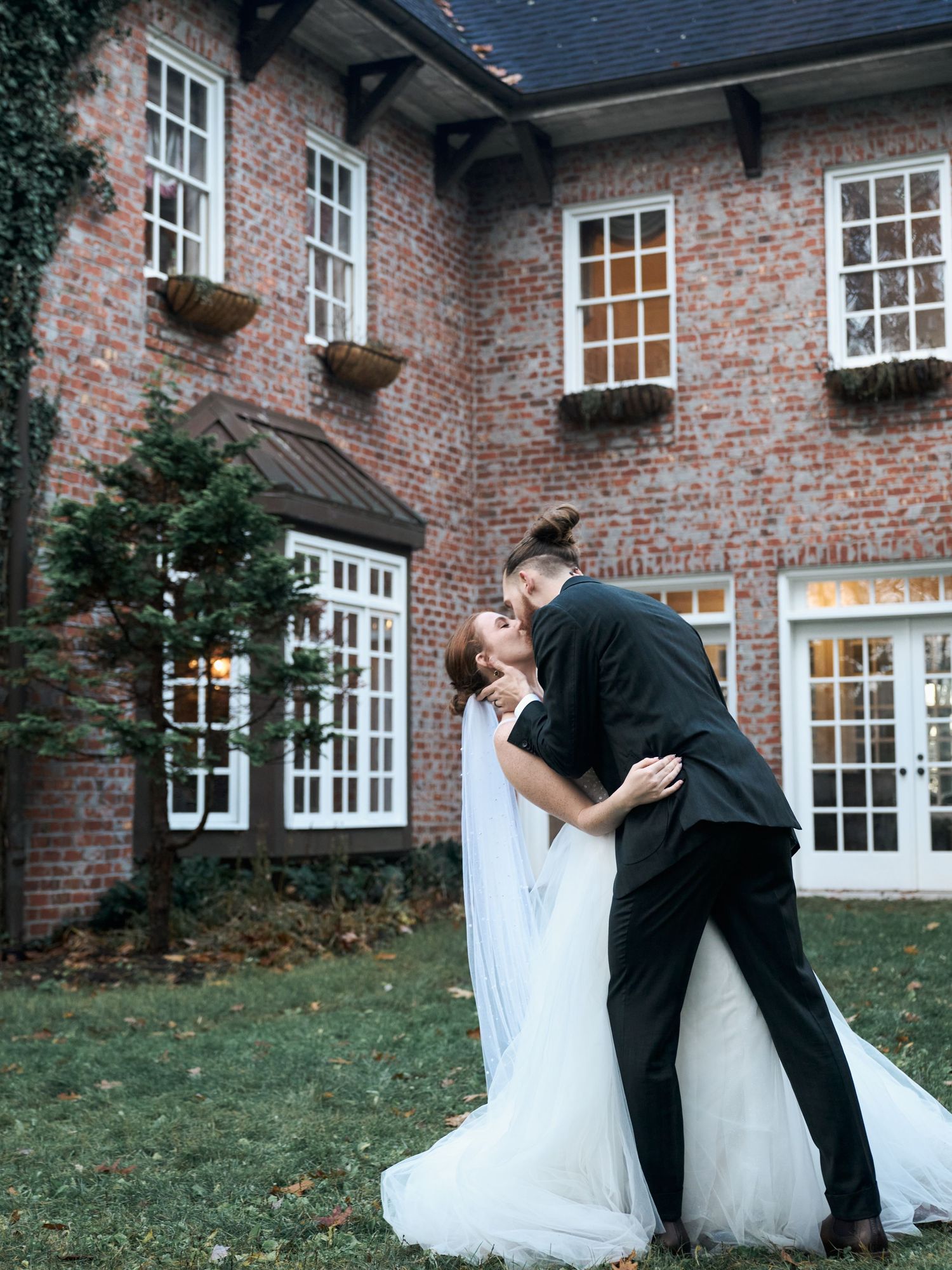 A romantic wedding moment in front of a traditional brick house with white-framed windows and ivy.