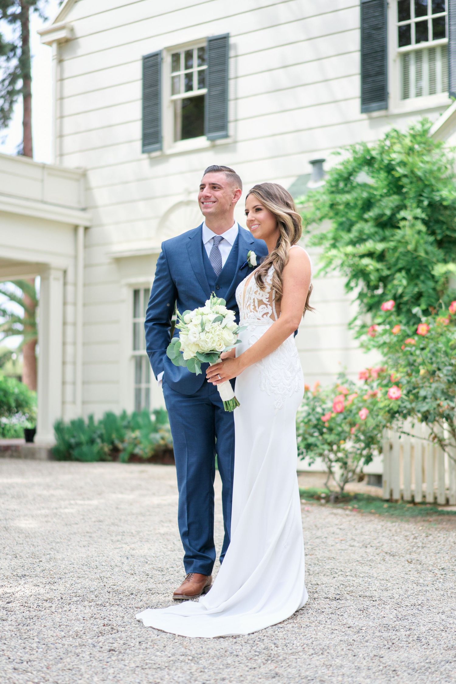 Newlyweds pose in front of a white colonial house with roses in bloom.