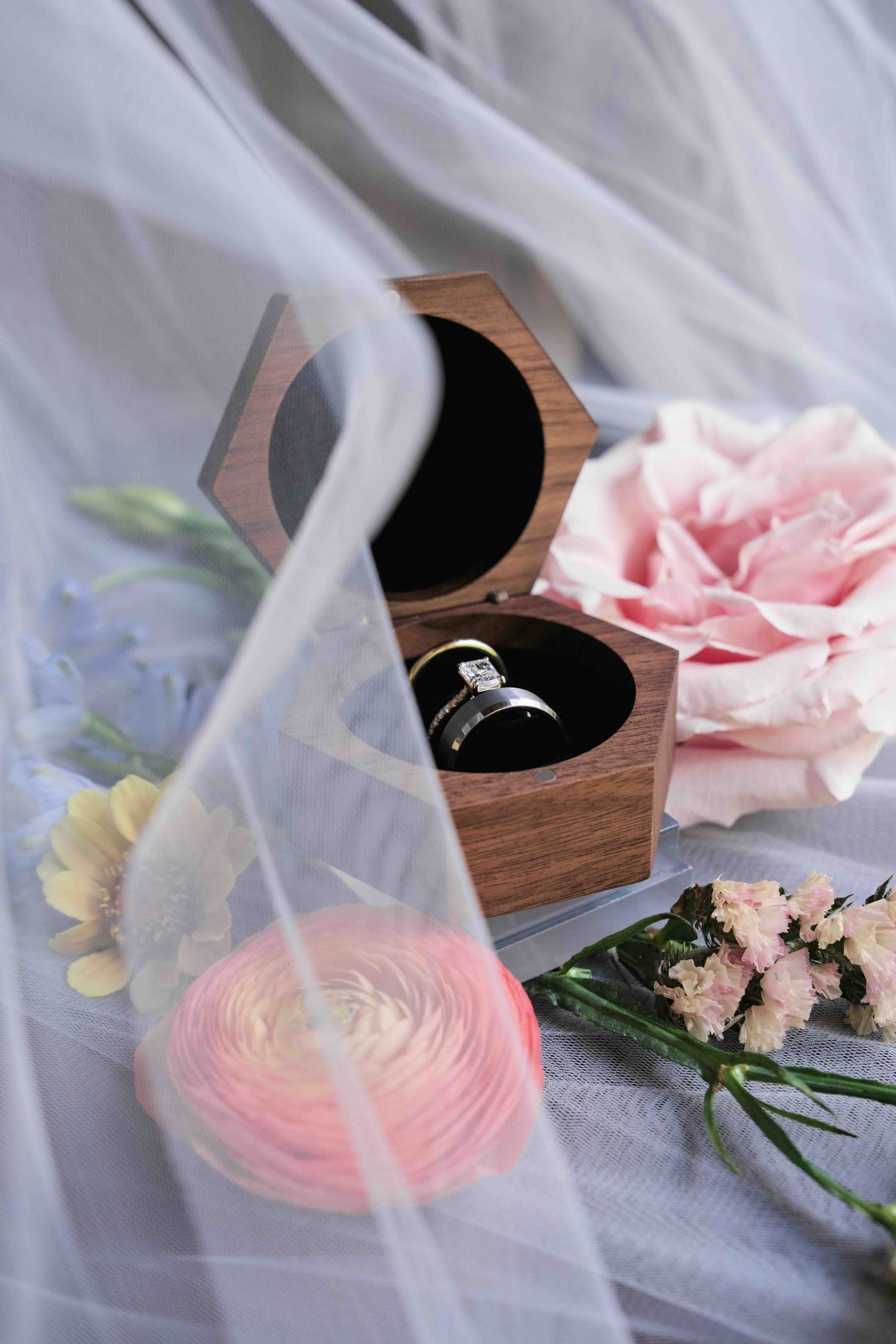 Elegant wedding ring in wooden box surrounded by pink roses and wildflowers under sheer bridal veil.