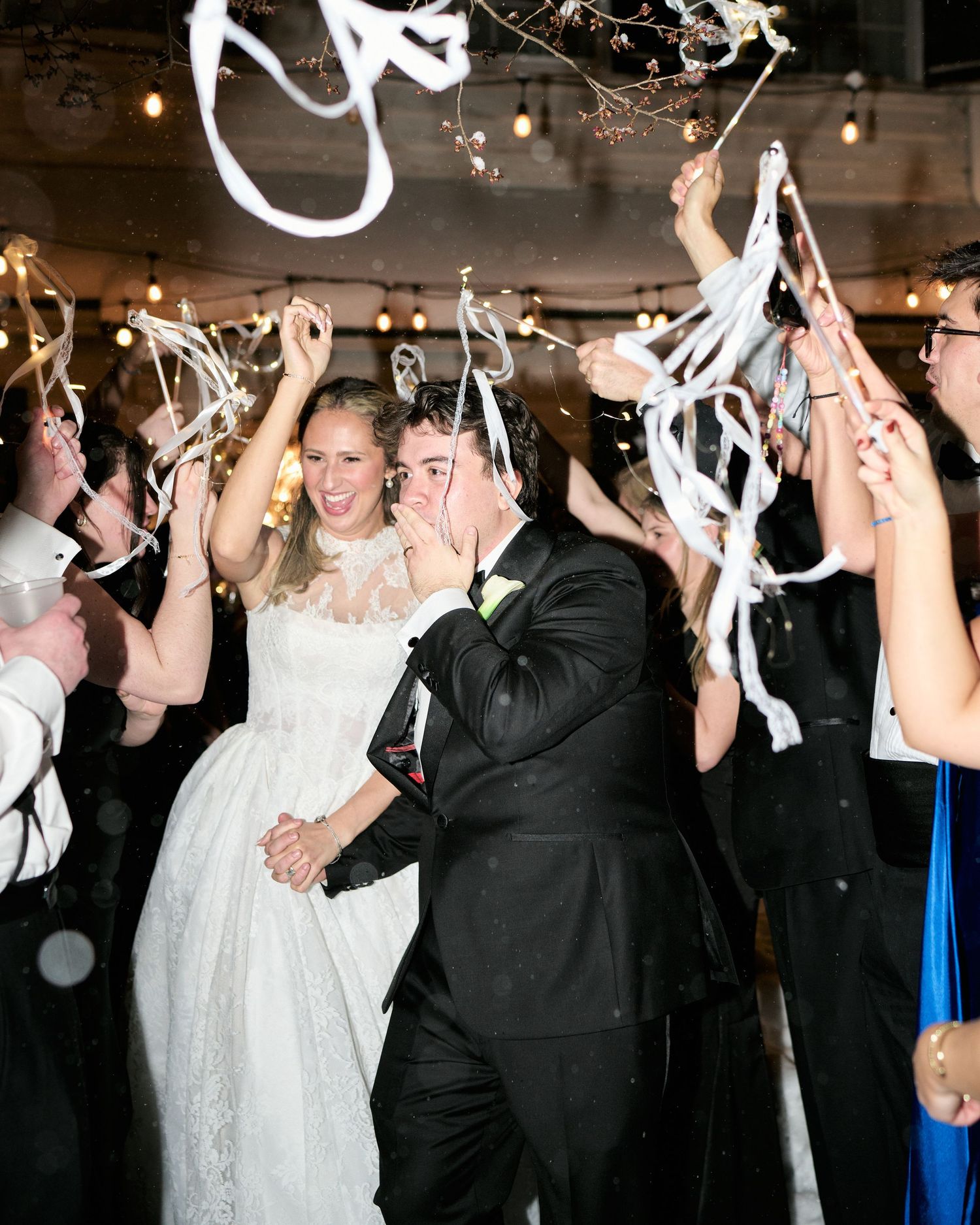 Bride and groom exit wedding reception through cheering guests waving white streamers in elegant venue.