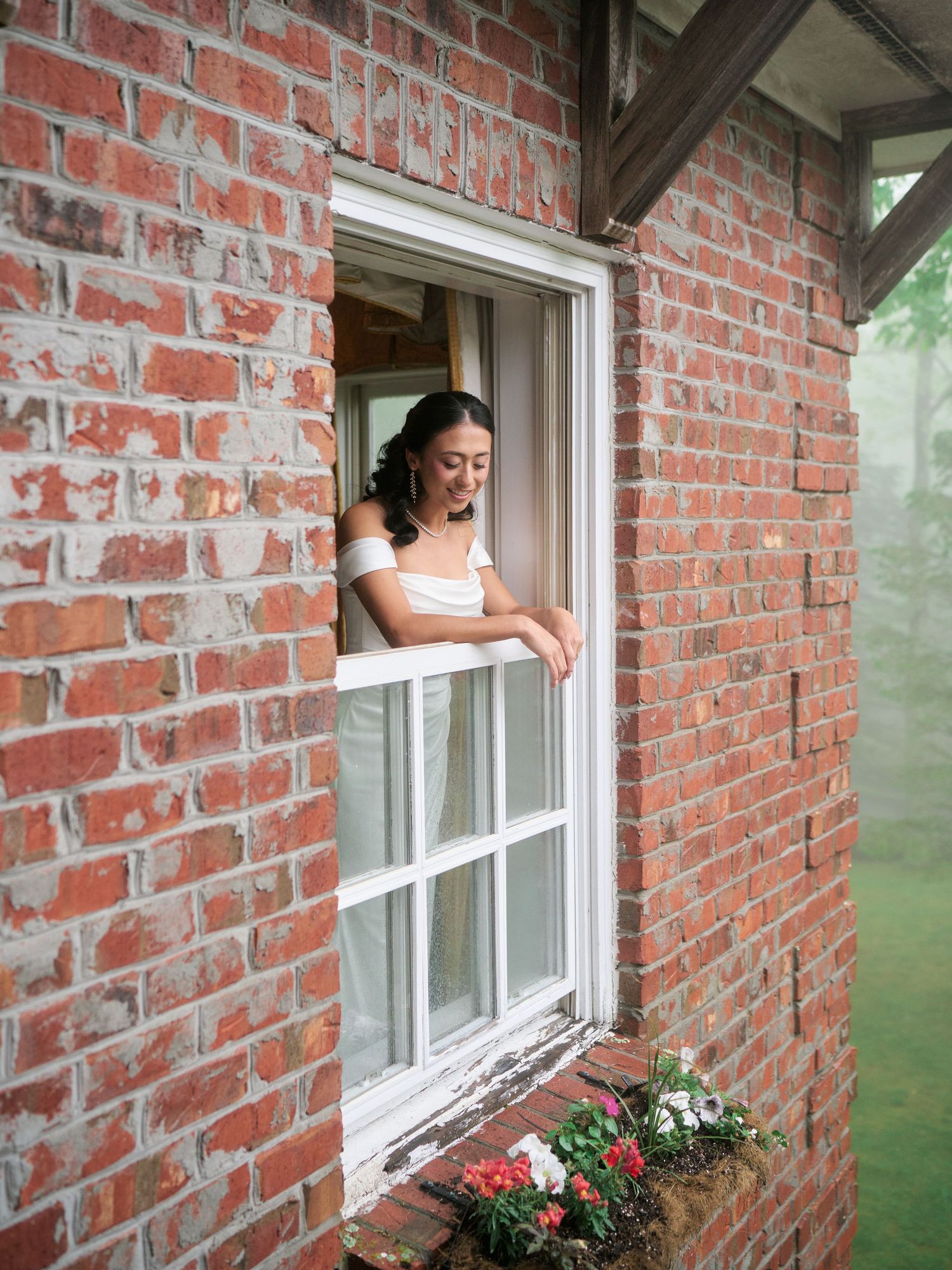Bride in white dress leaning out of a brick building window with flower box below.