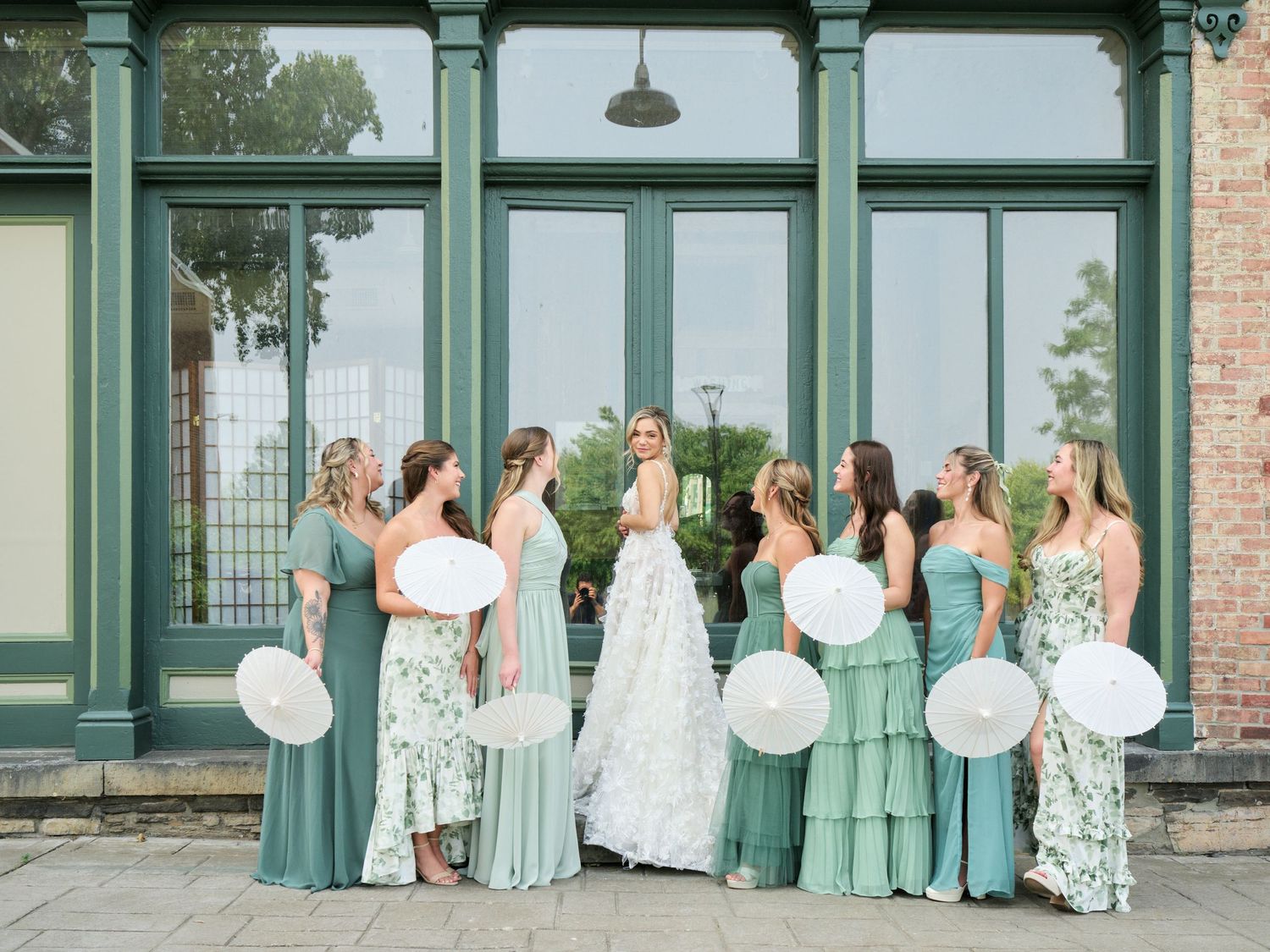 A group in coordinating mint green and floral dresses pose together holding white paper lanterns outside a building.