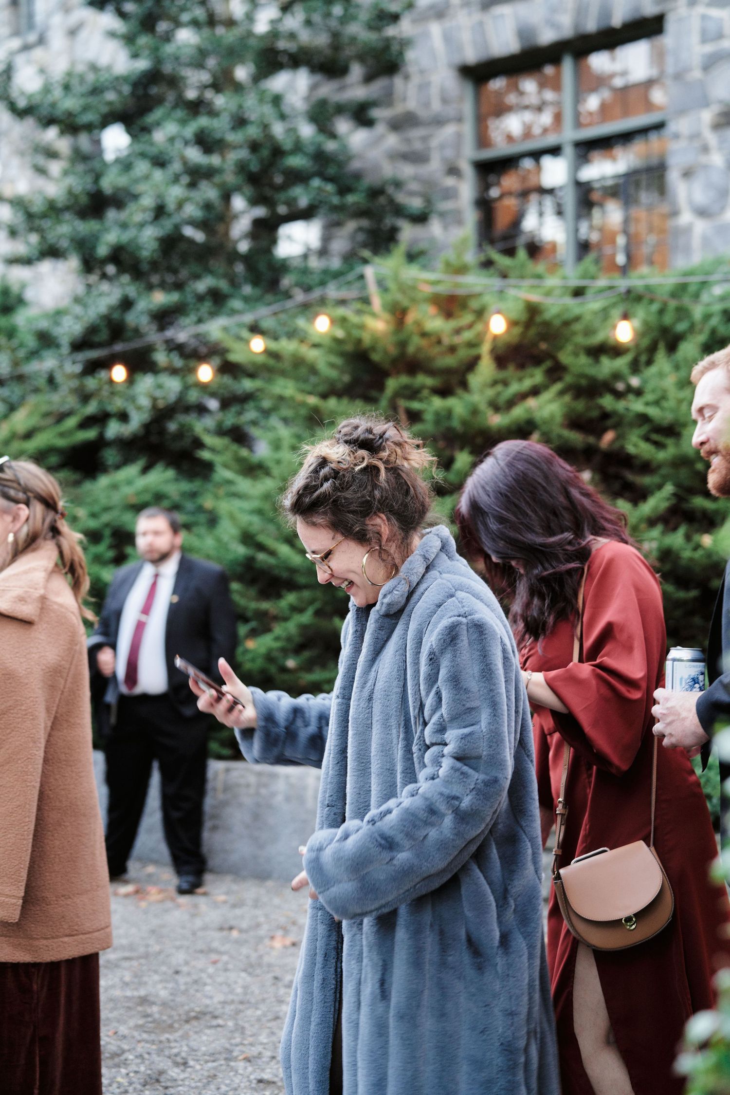 Wedding guests in winter coats checking their phones outside a stone building with string lights.