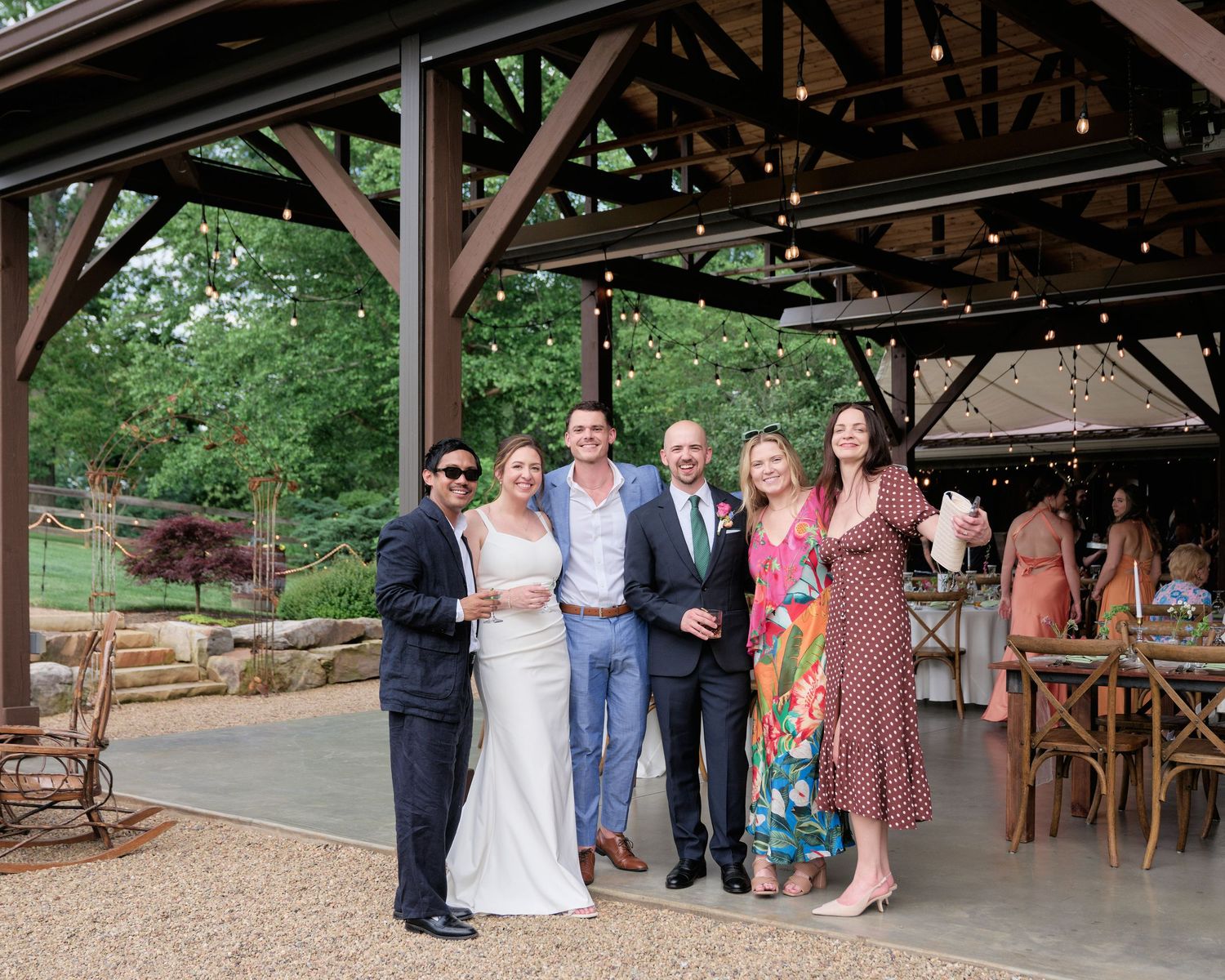Wedding party poses under a rustic wooden pavilion at outdoor venue.