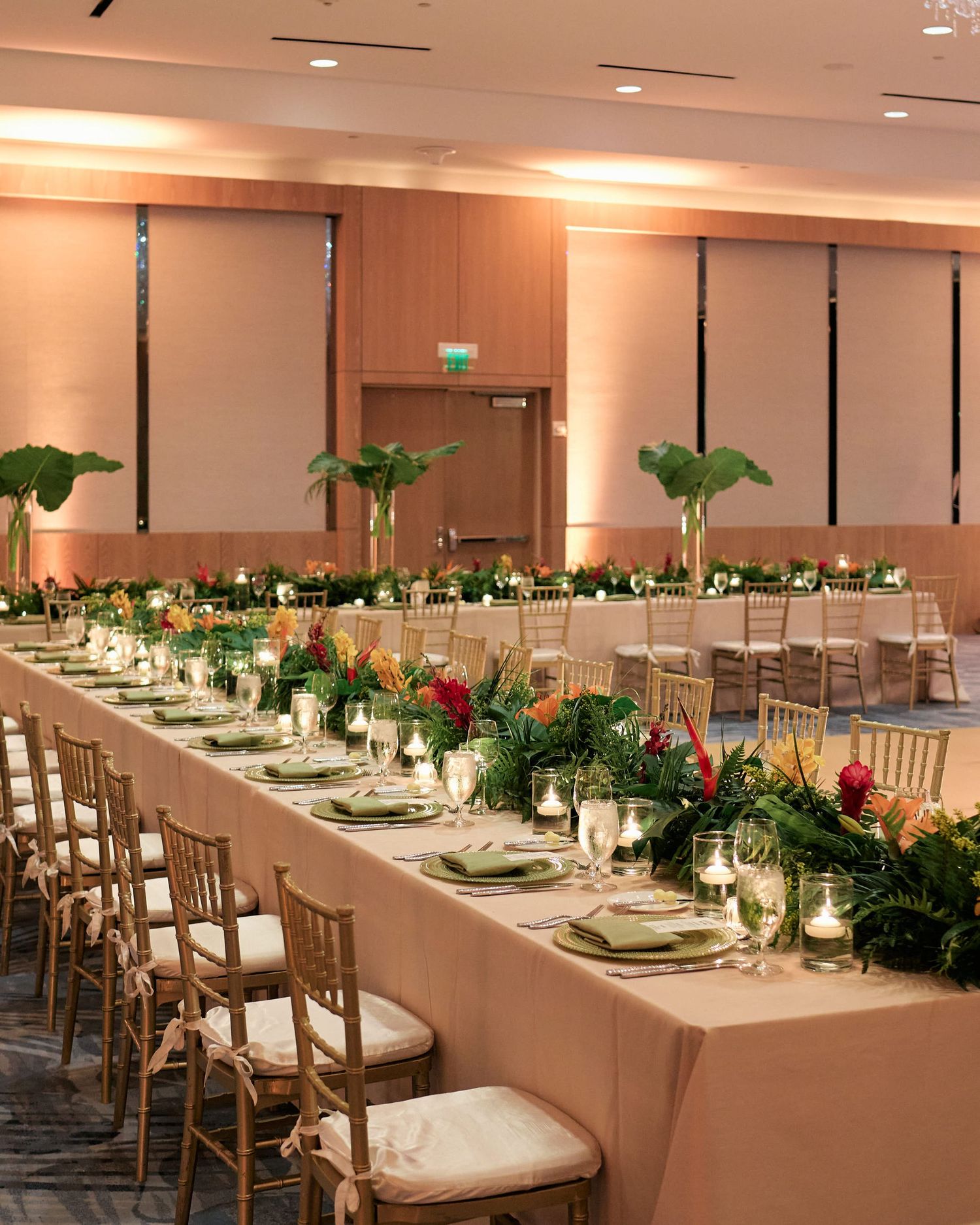 Elegant wedding banquet table with tropical greenery garland runner and golden chairs in a modern event space.