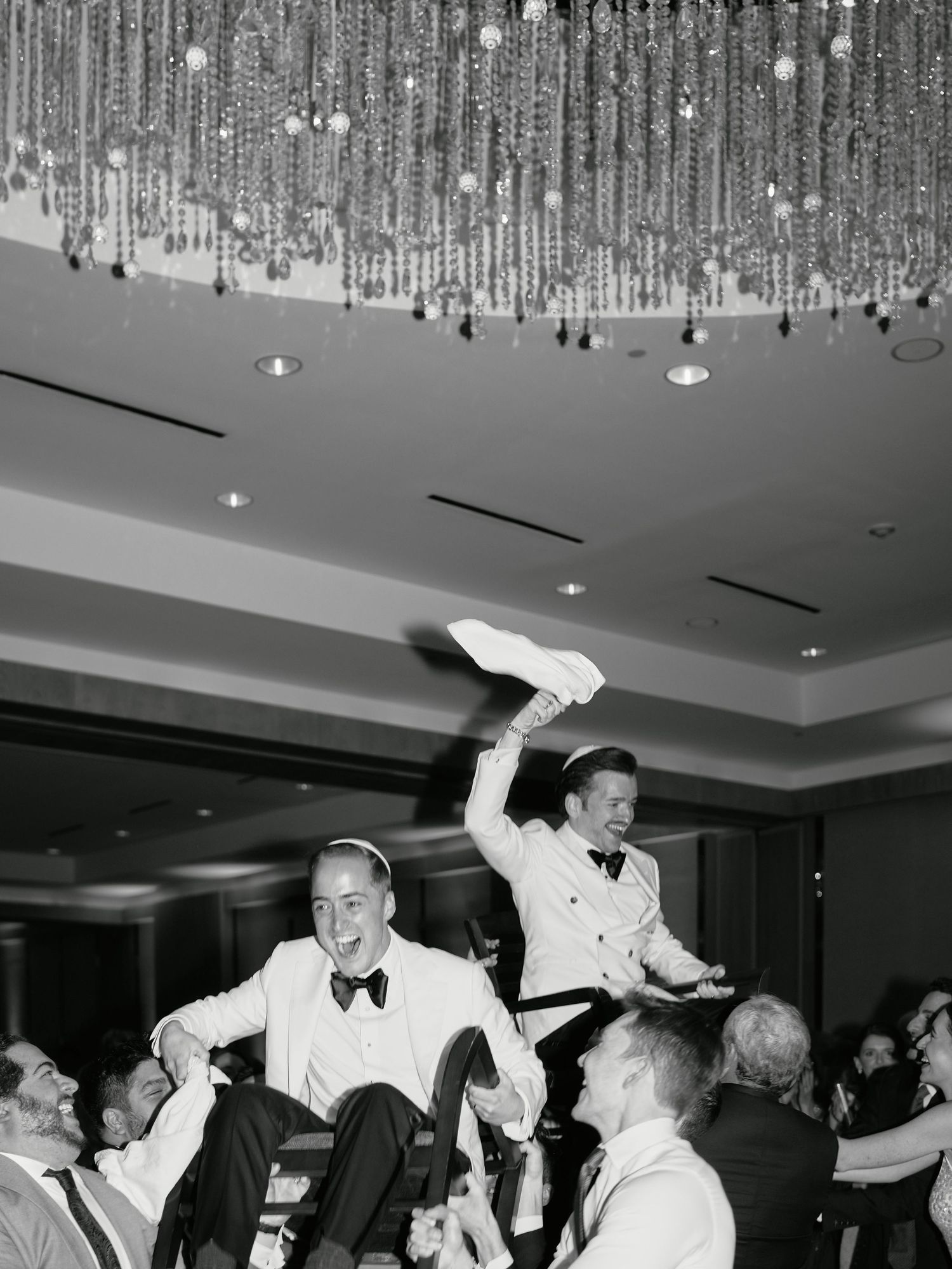 A vintage black and white photo shows two people being lifted up at a lively formal party with crystal decorations.