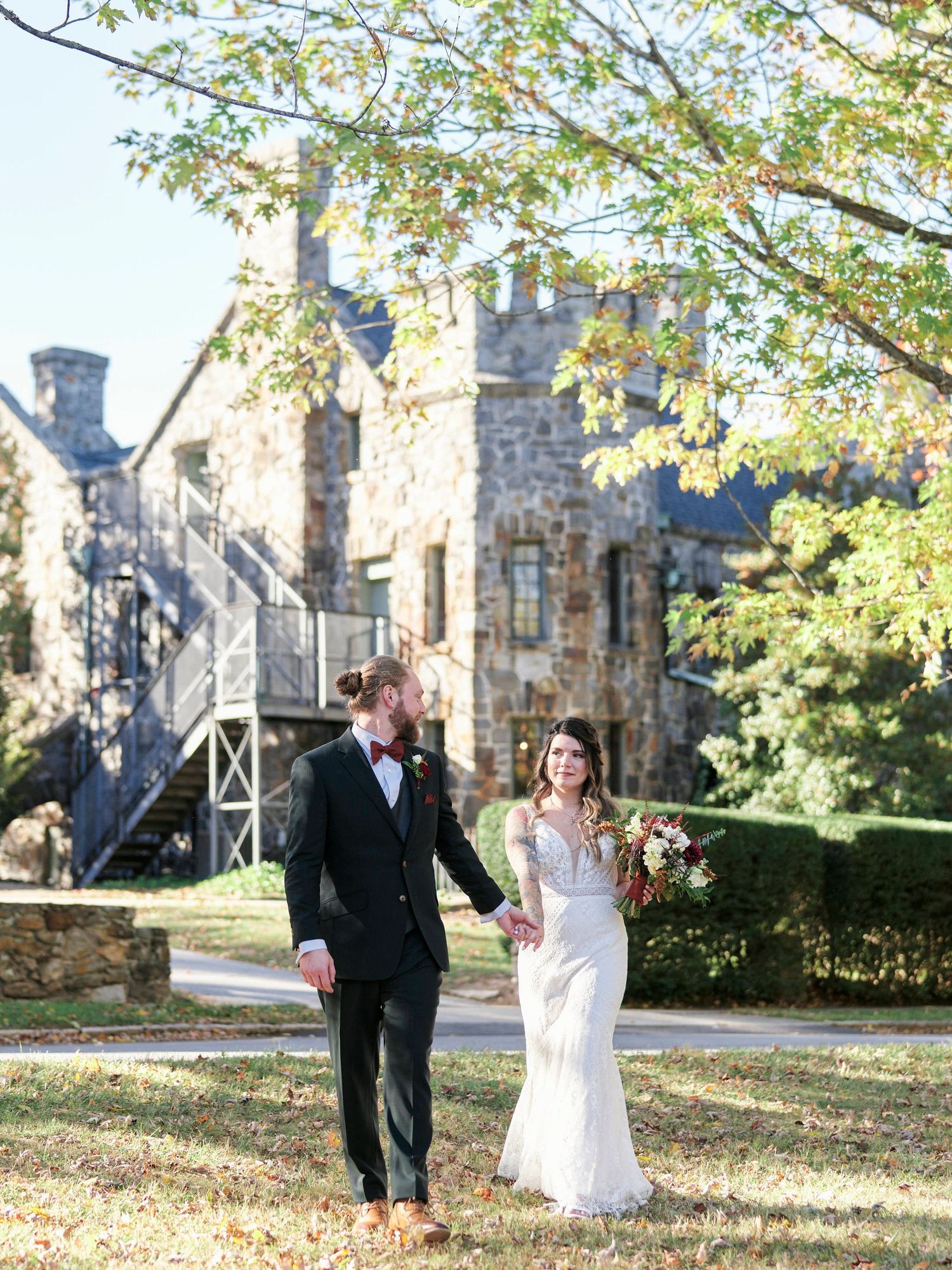 Bride and groom walking hand in hand in front of stone castle on sunny day.