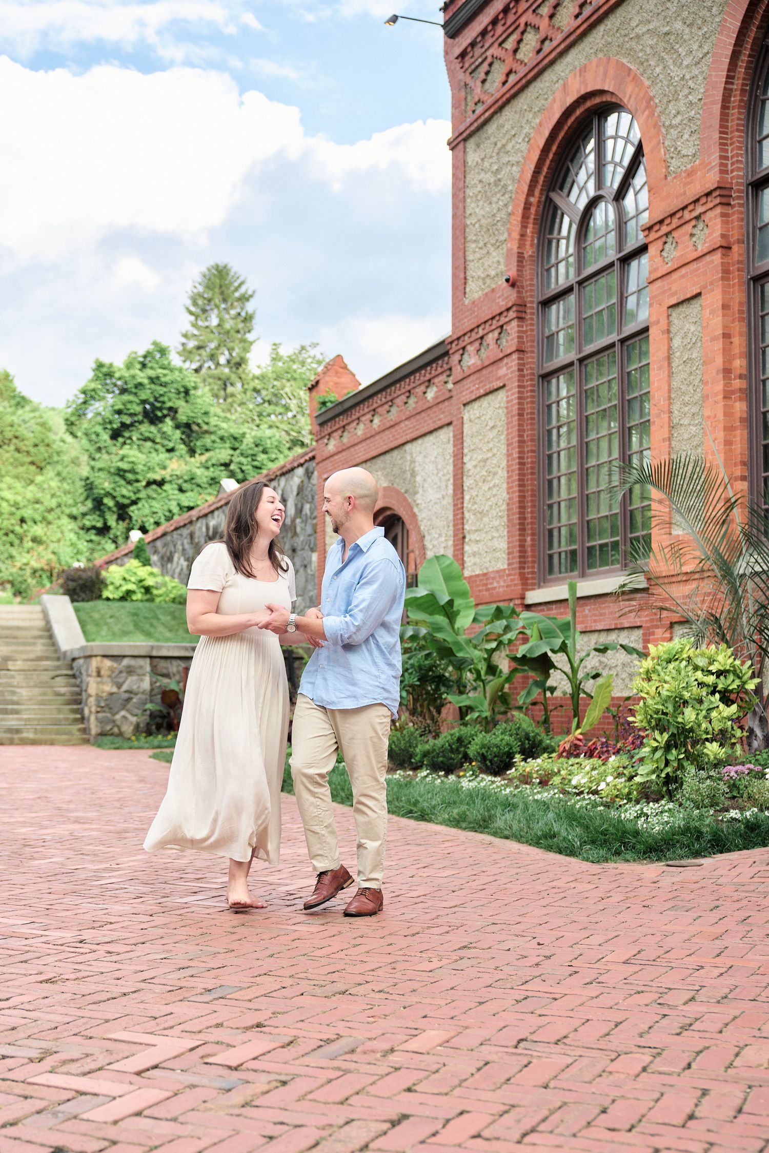 A couple shares a joyful moment outside a brick conservatory with arched windows.